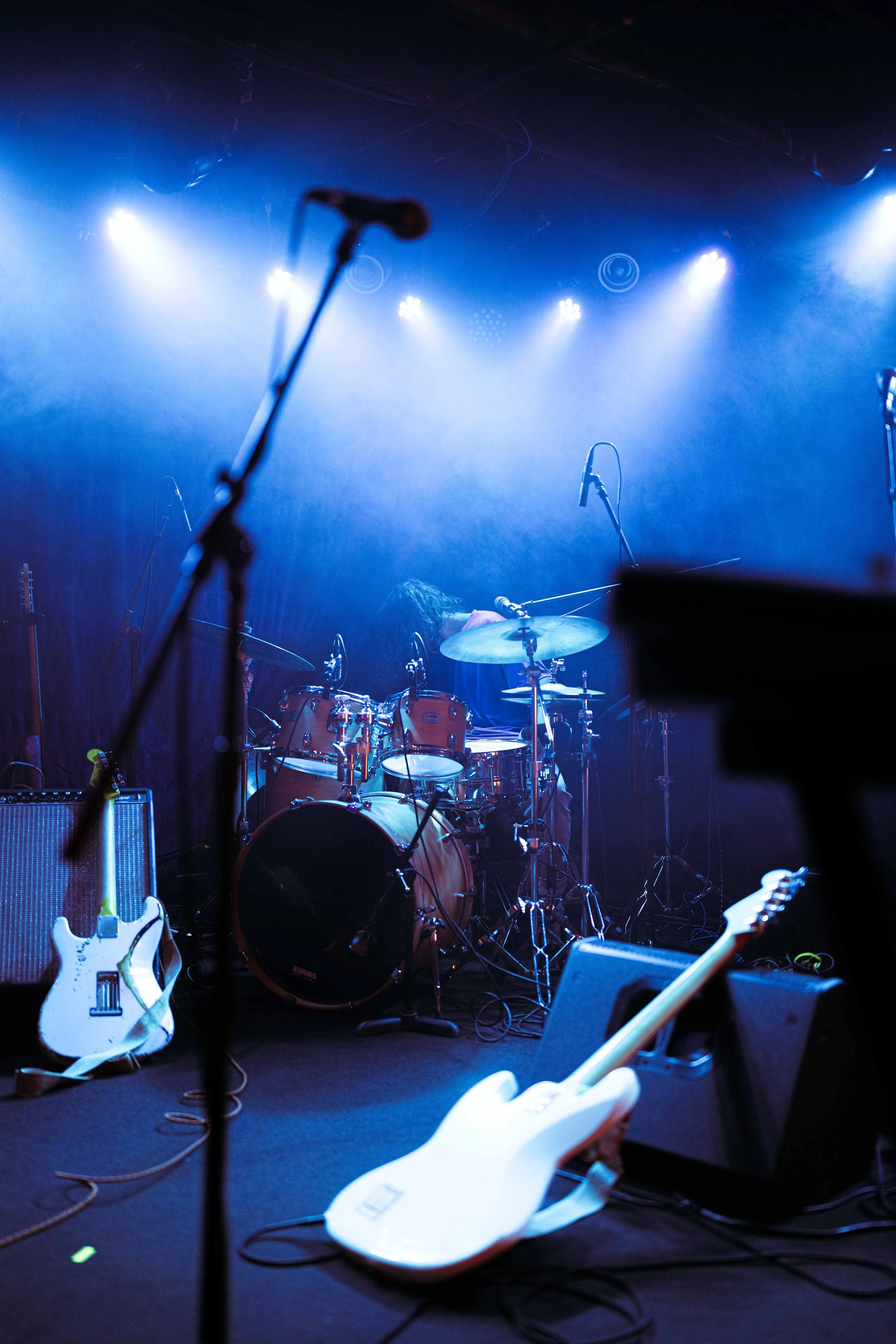 Empty stage with musical instruments, including electric guitars, a drum set, and amplifiers, illuminated by blue stage lighting.