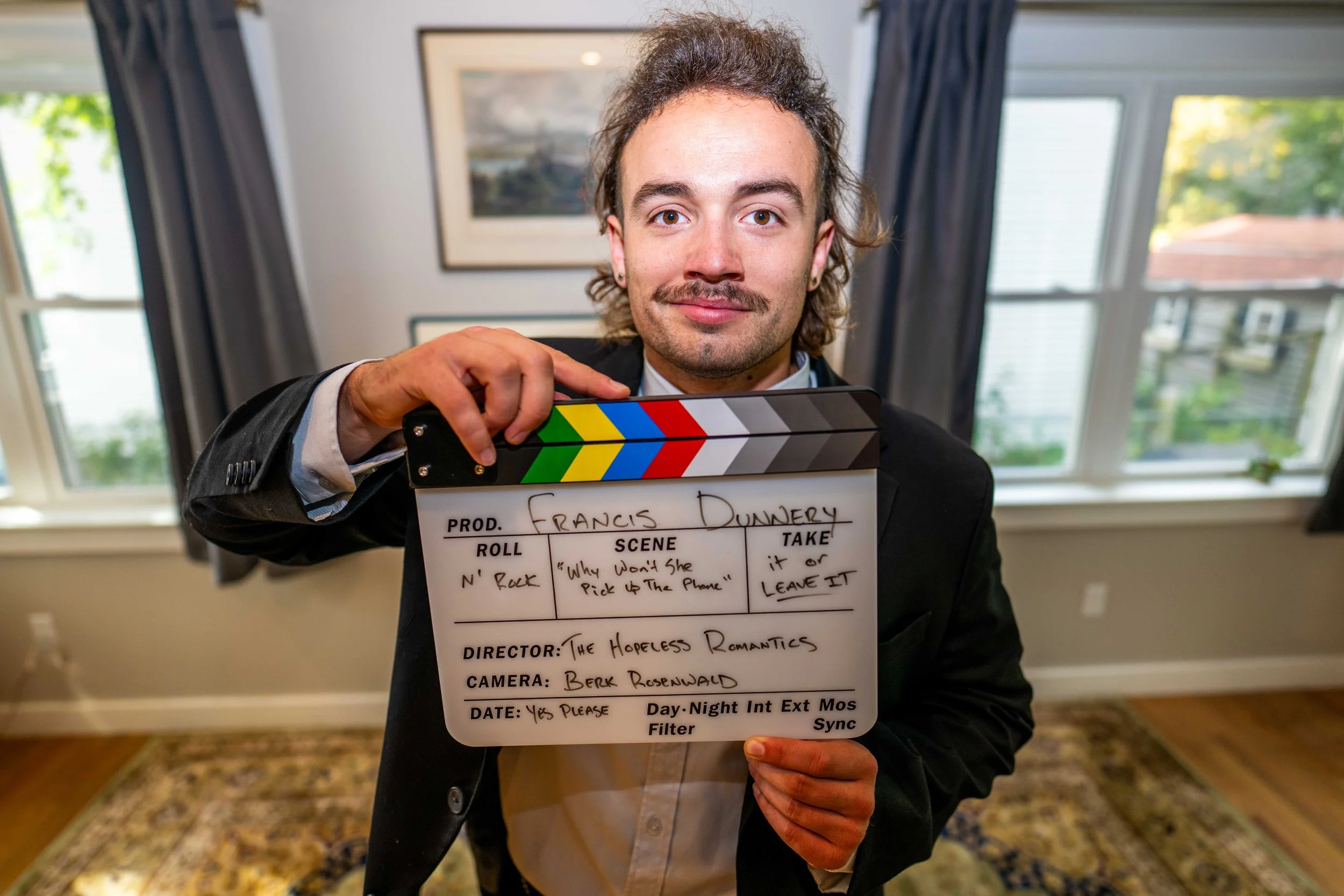 Man in formal suit holding a movie clapperboard in a well-lit room with large windows.