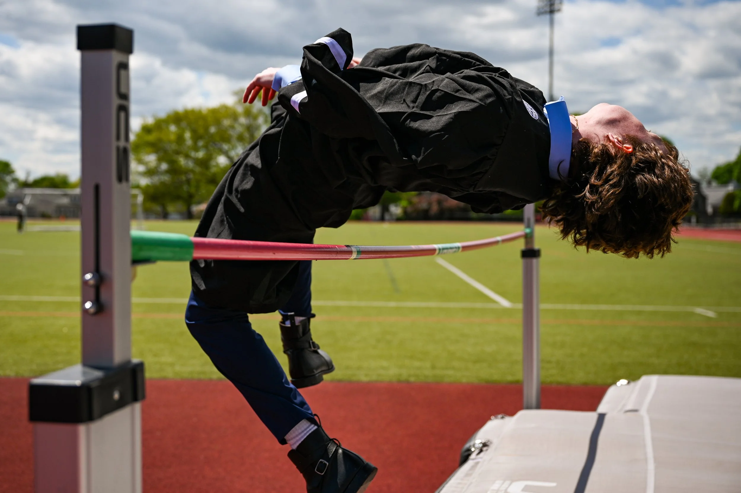 Student in a black gown and blue pants attempting a high jump over a red and green bar during outdoor track and field practice on a grassy and track field under cloudy sky.