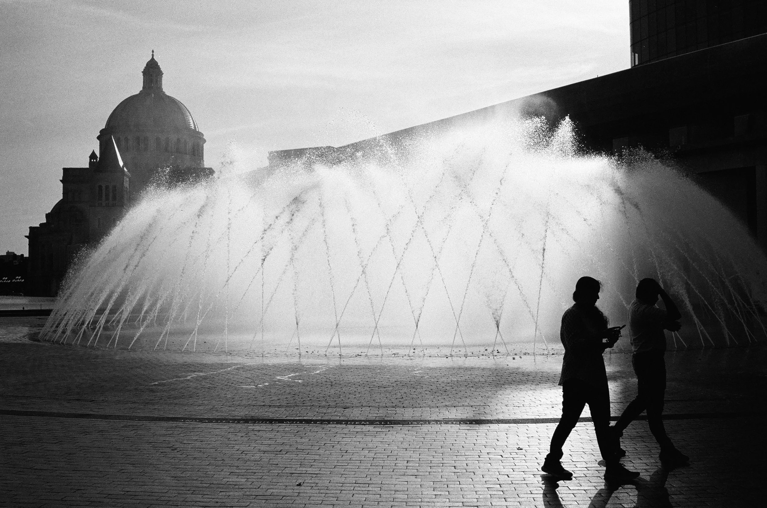Silhouettes of two people walking past a large fountain with water spraying in front of a historical building with a domed roof.