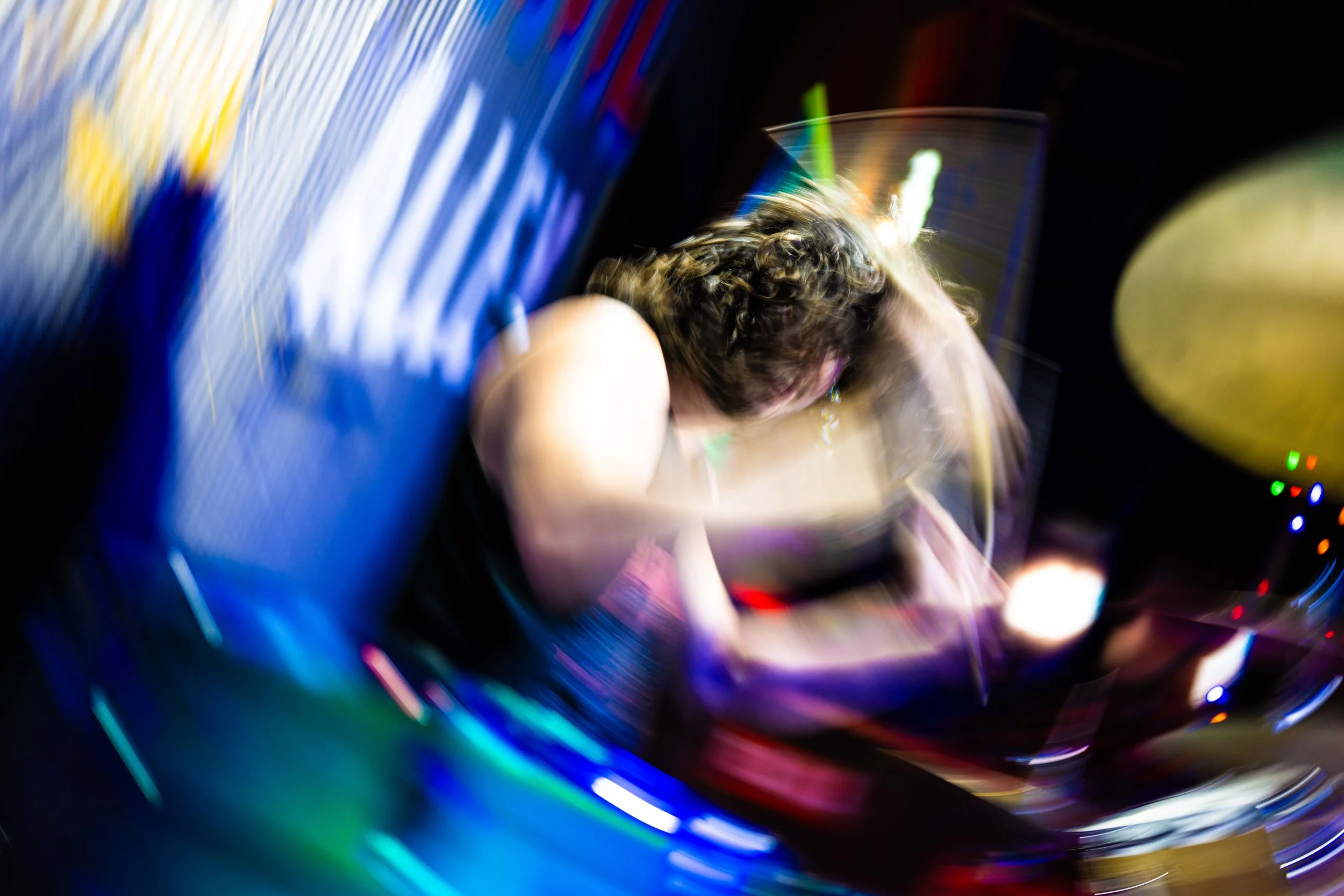 A person with curly hair is sitting at a table, holding a glass of drink, with colorful, blurred lights and reflections around.