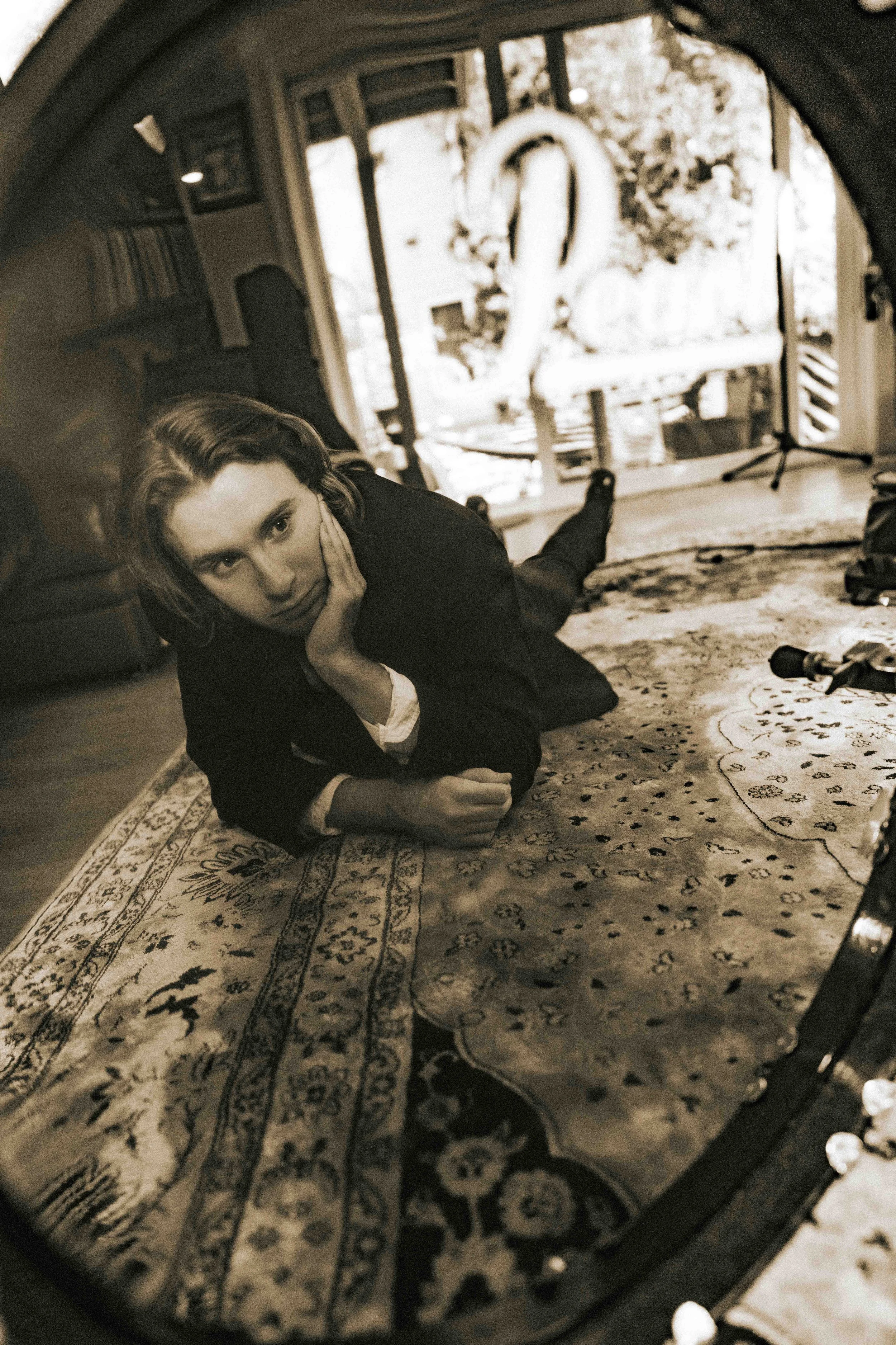 A woman lies on her stomach on a patterned rug, resting her head on her hand, looking at a mirror that reflects her face and the room behind her.