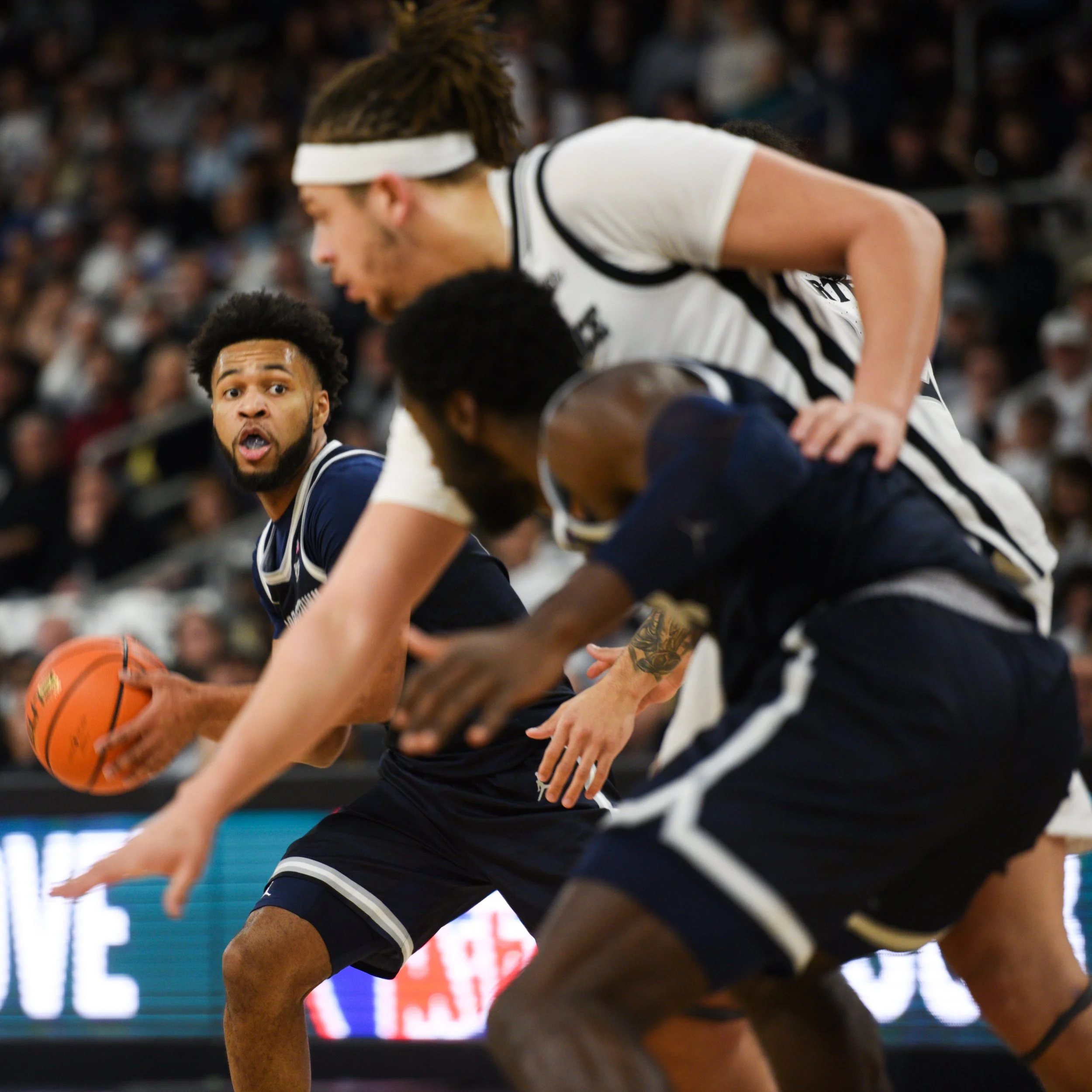 Three basketball players in action during a game, with one player holding the ball while the others defend