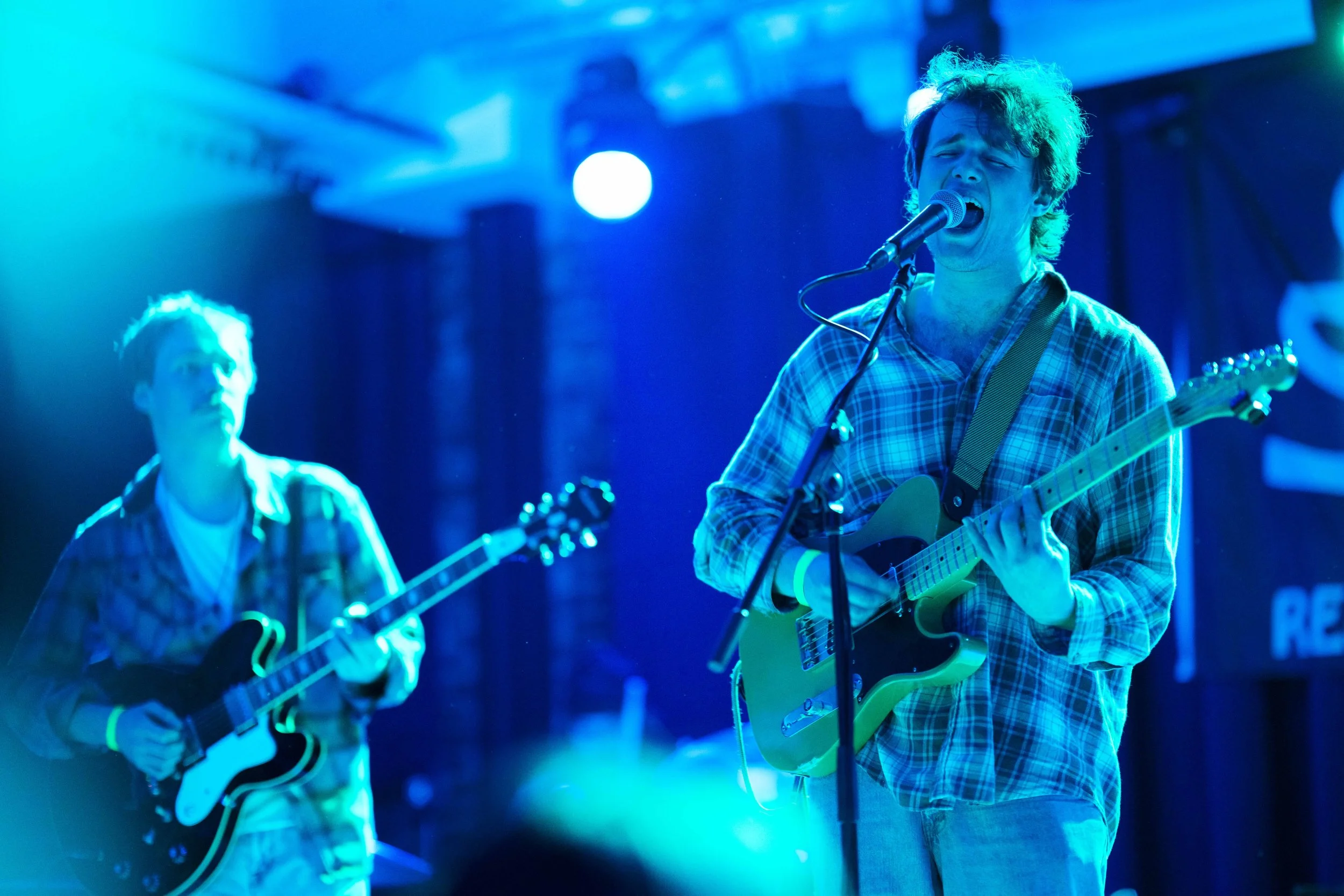 Two young men performing on stage with guitars, one singing into a microphone, illuminated by blue stage lighting.