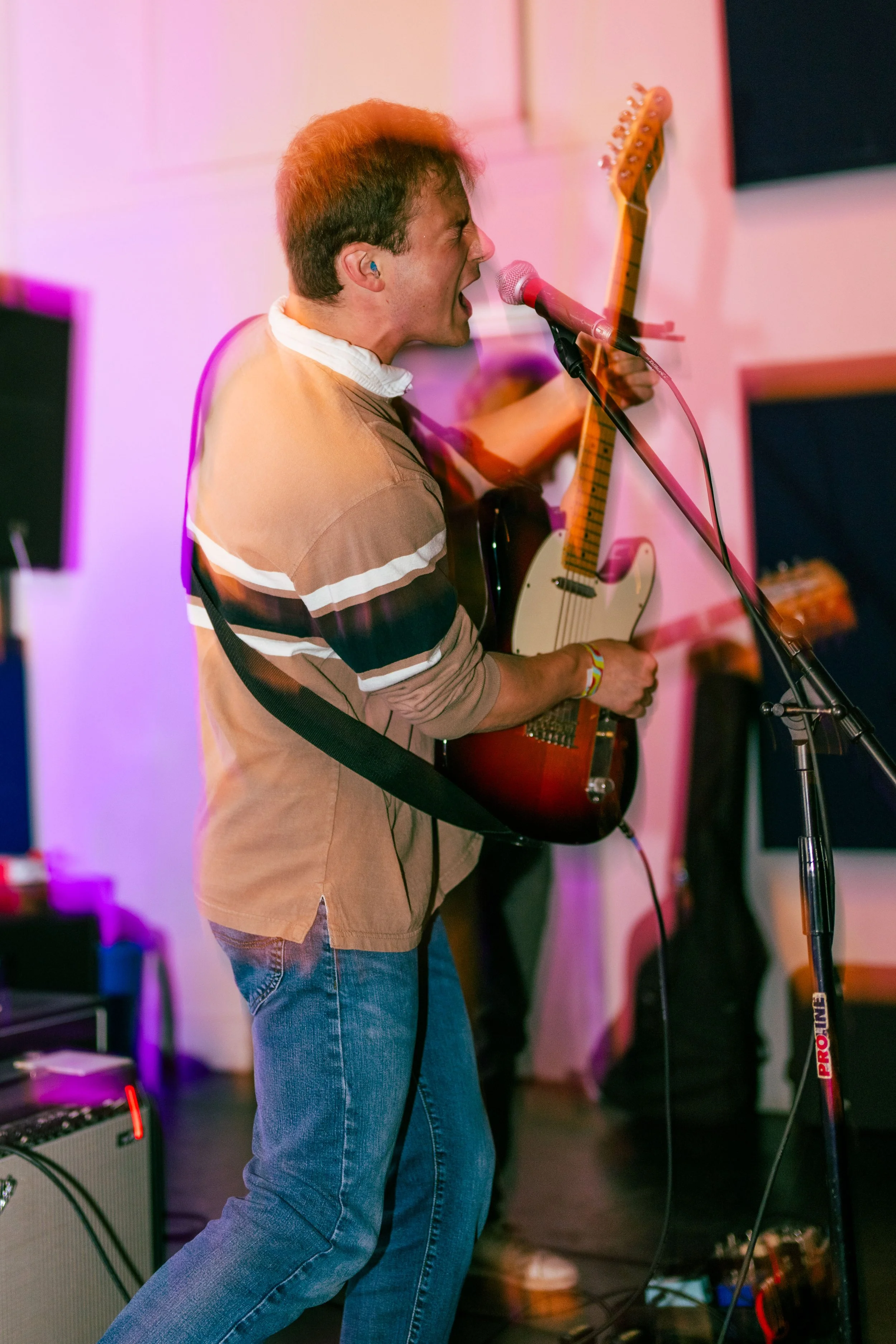 A man singing and playing an electric guitar into a microphone during a live music performance in a room with pink lighting.