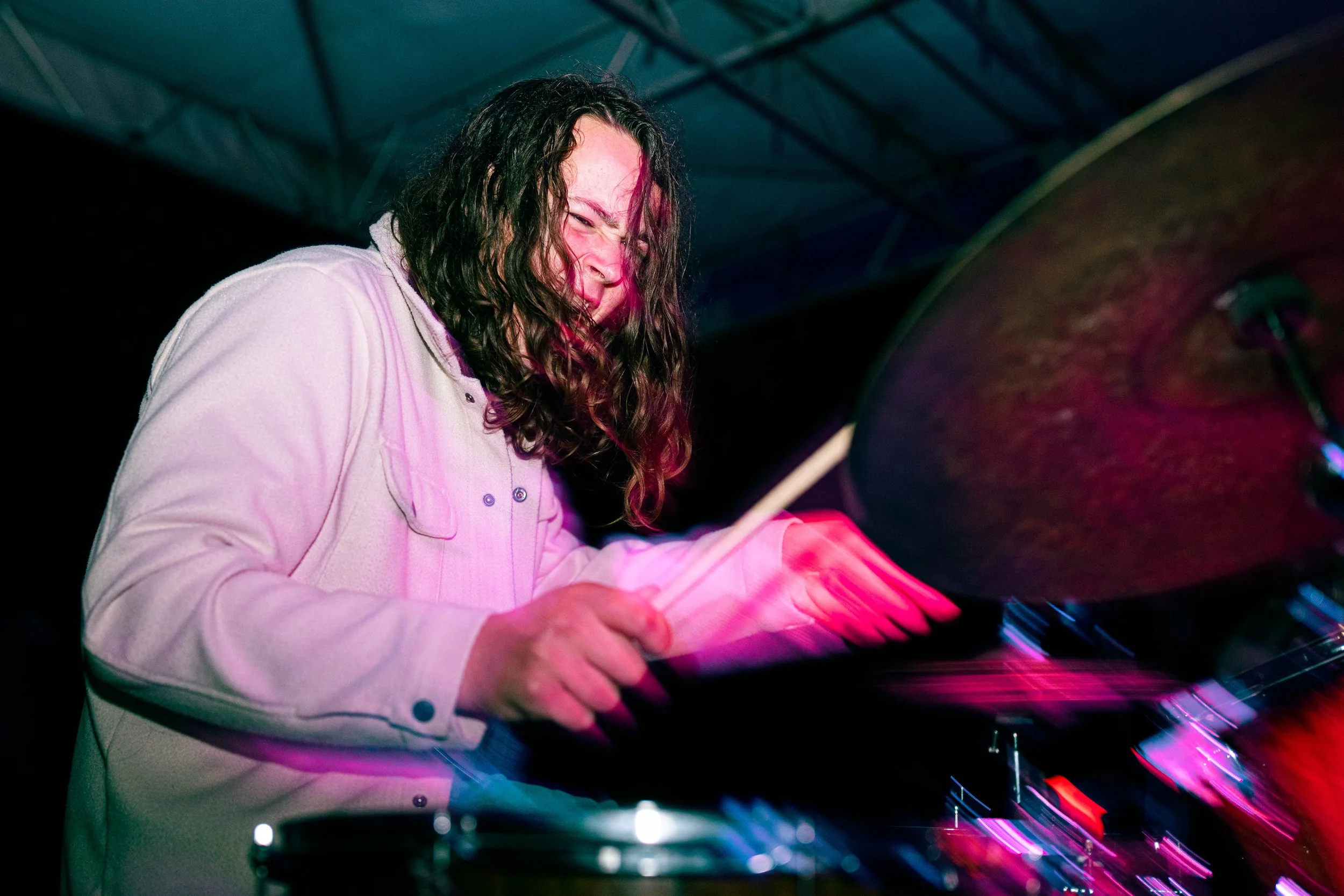 A person with long dark curly hair playing drums with a passionate expression, under colorful stage lighting.
