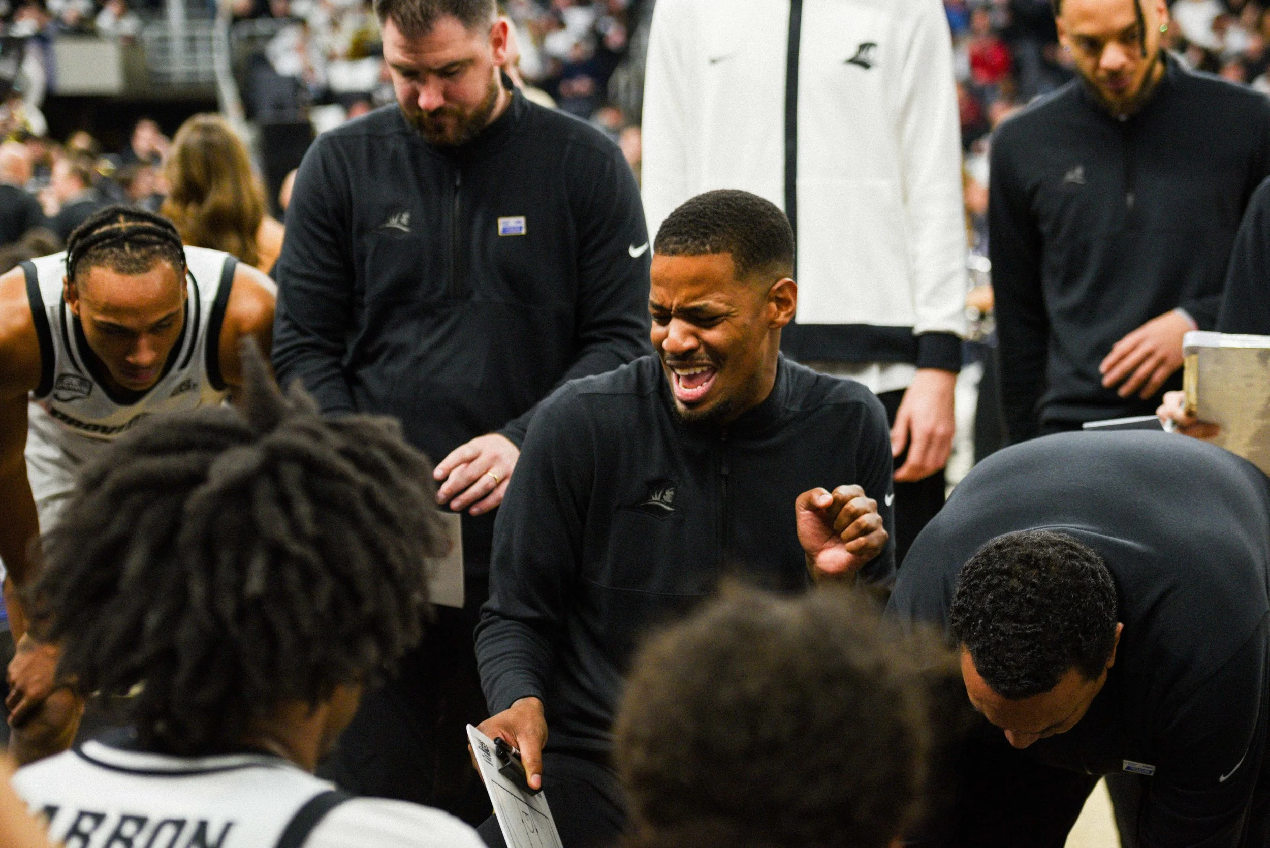 A basketball coach passionately coaching his team suggests an intense moment during the game. The players are gathered around him, listening attentively, in a busy arena with an audience in the background.