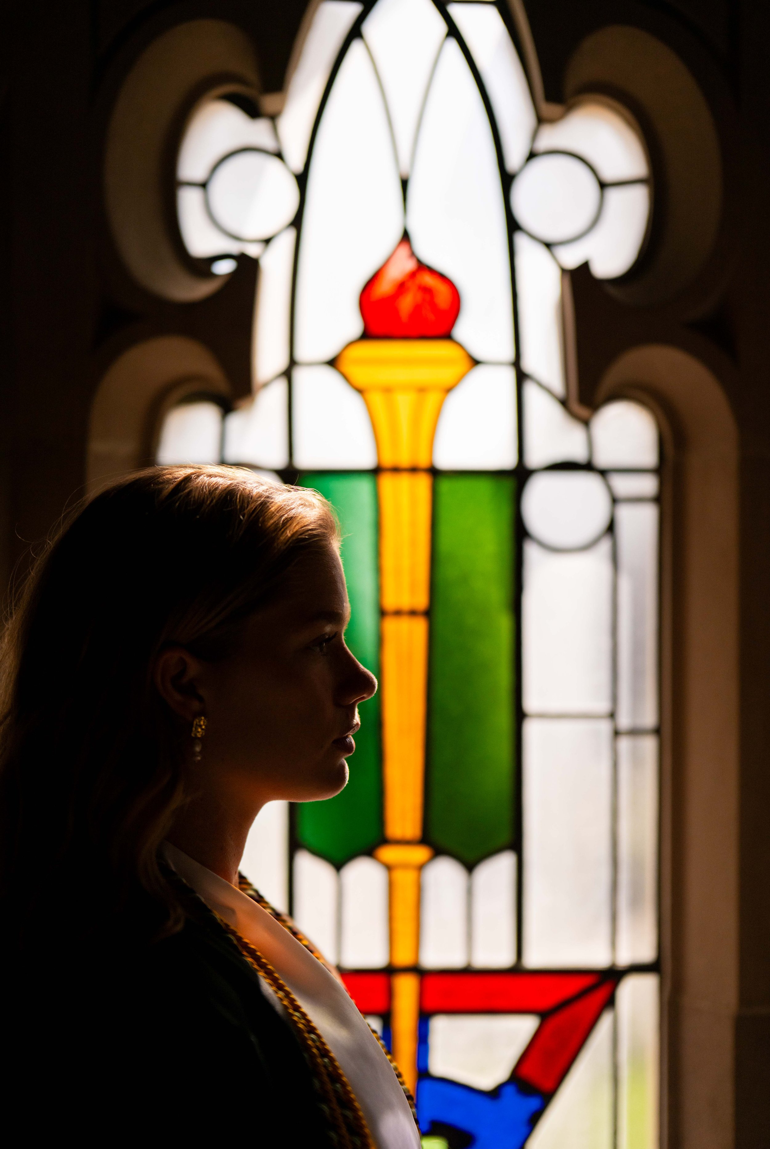 Profile of a woman with blond hair and earrings standing in front of a colorful stained glass window.
