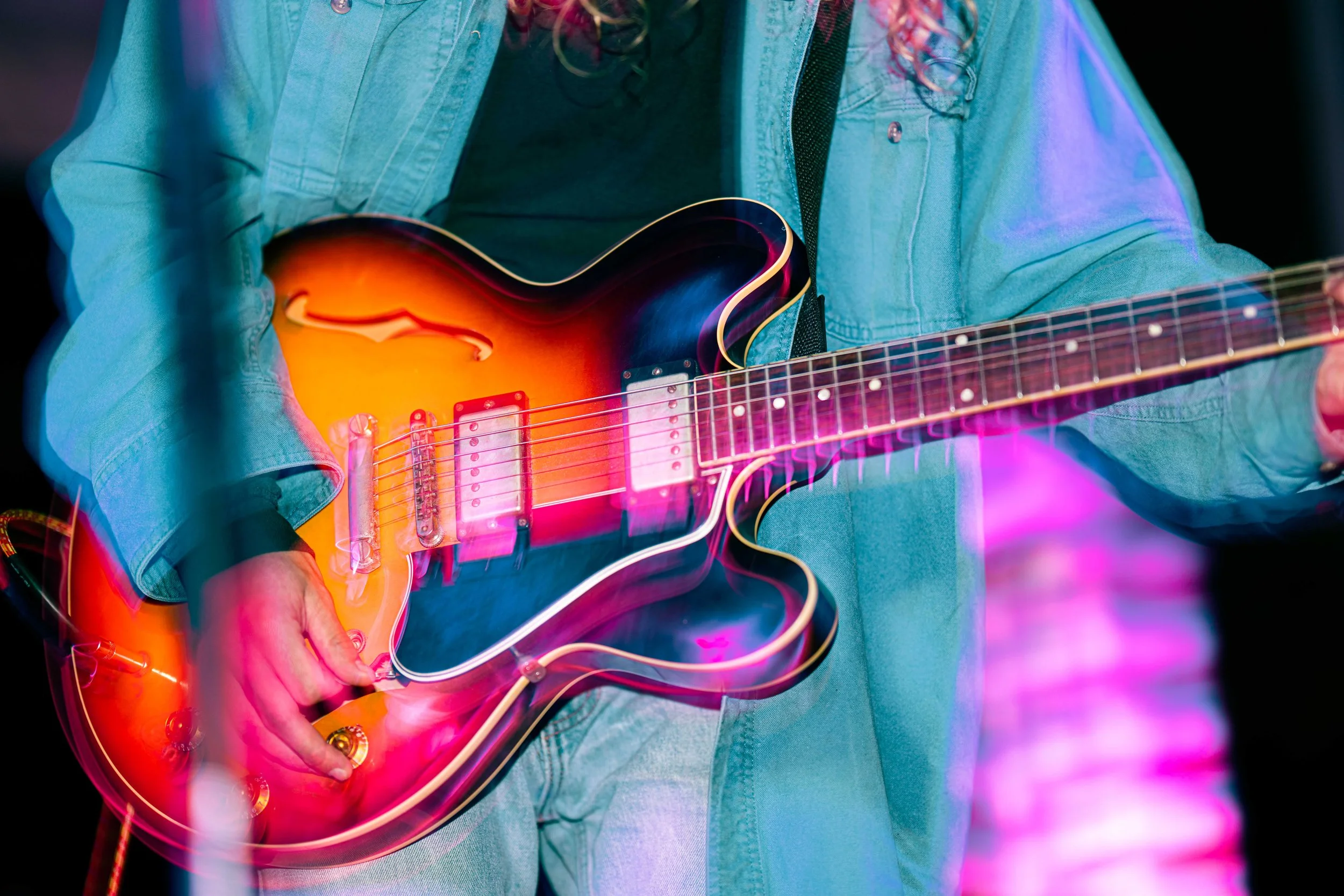 Person wearing a green jacket playing an orange sunburst electric guitar with pink lighting effects.