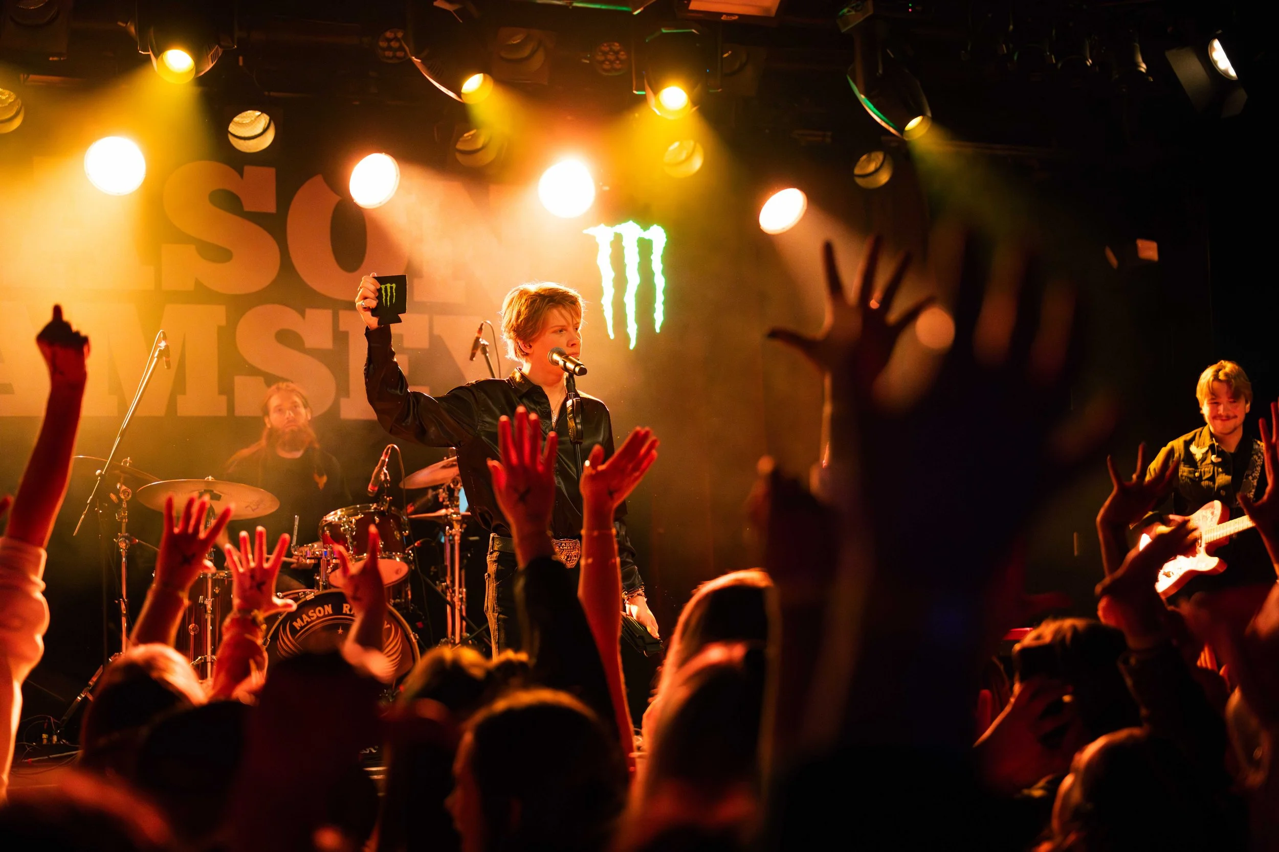 A live music performance with a band on stage, including a singer with a microphone holding a Monster energy drink, a drummer, and a guitarist. Audience members are raising their hands towards the stage, with stage lights illuminating the scene and a
