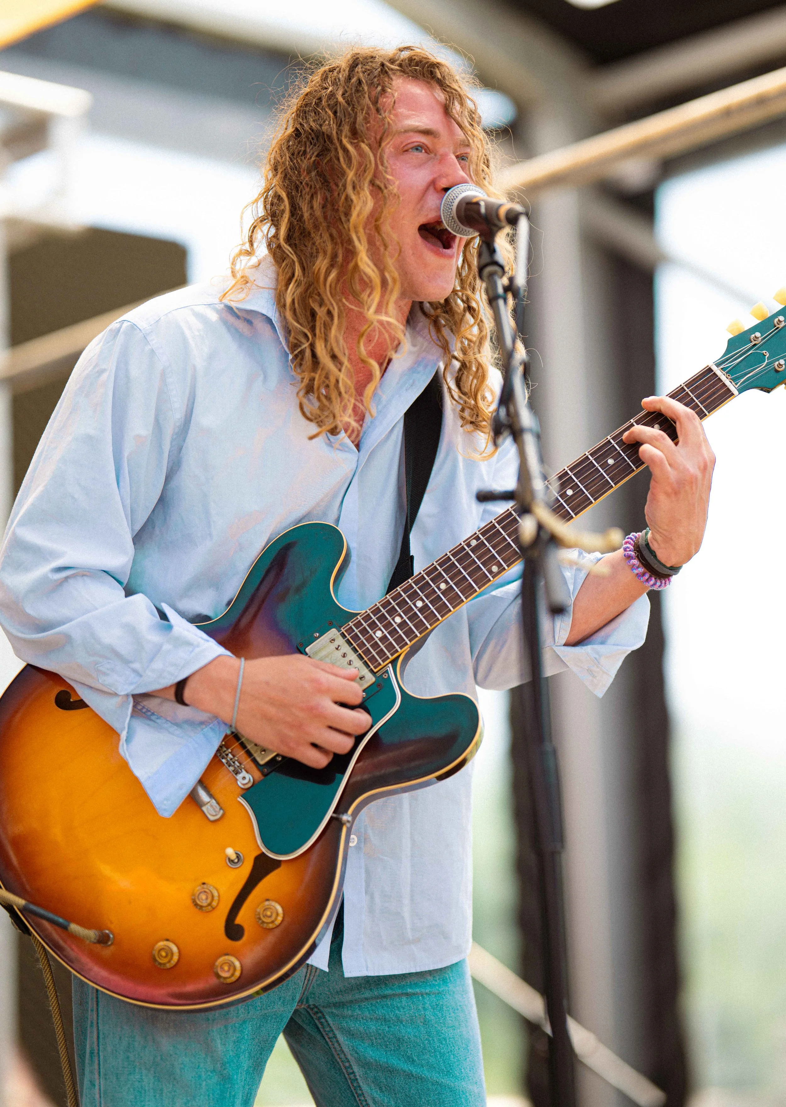 A person with long curly hair singing into a microphone while playing an electric guitar on an outdoor stage.