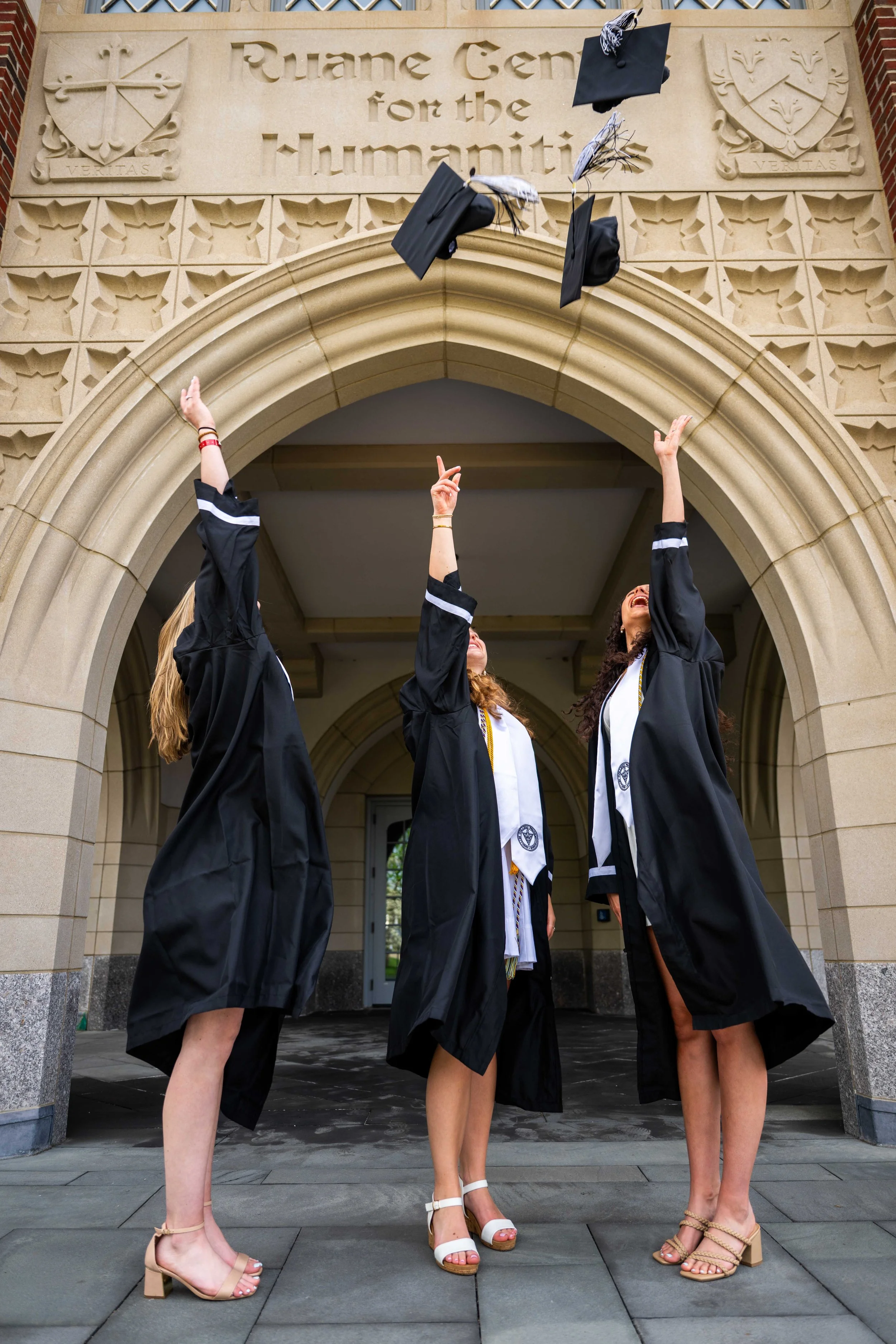 Three graduates in caps and gowns tossing their caps into the air outside a building with an archway that reads "Ruan Center for the Humanities."