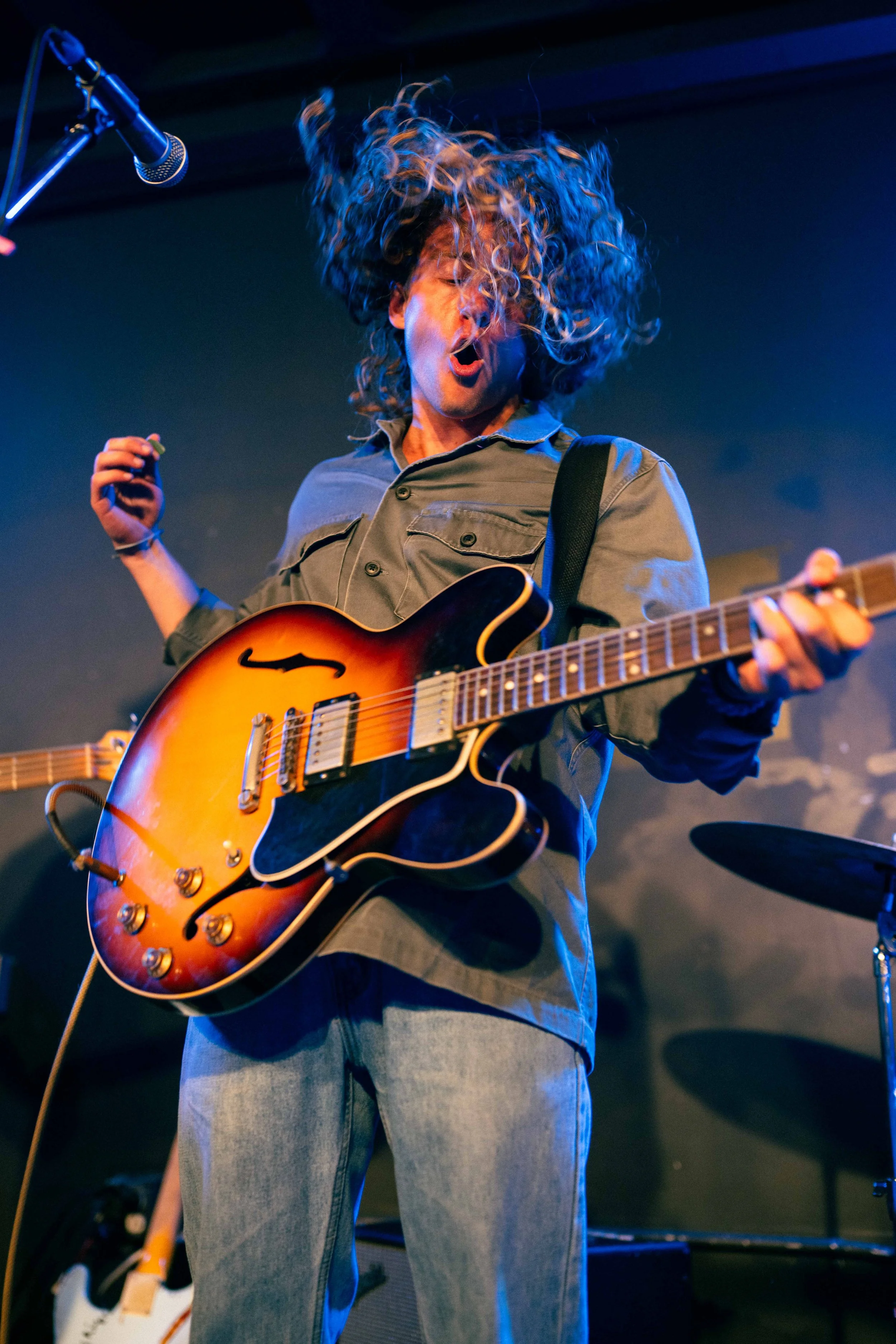 A musician with curly hair passionately performs on stage, playing an electric guitar with a sunburst finish, in front of a dark background with stage lights.