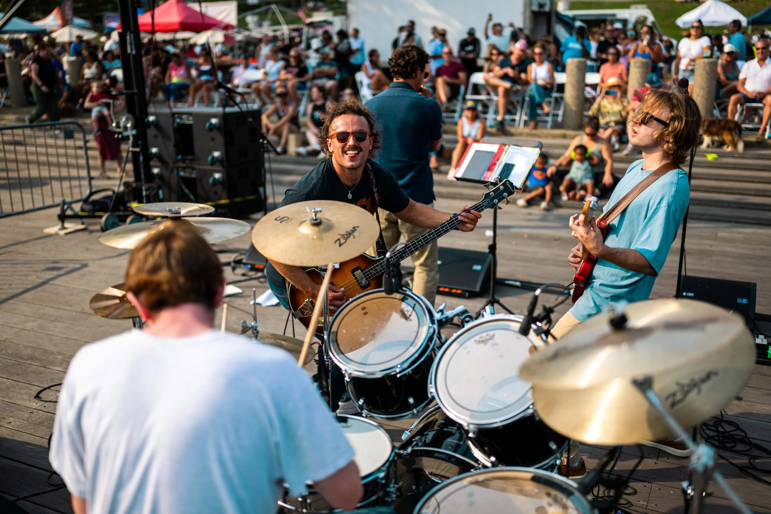 A band of four musicians performing outdoors on a wooden stage, with a drummer in the foreground, a guitarist with sunglasses smiling, another guitarist with long hair, and a bassist. A large audience is sitting and standing in the background, watchi