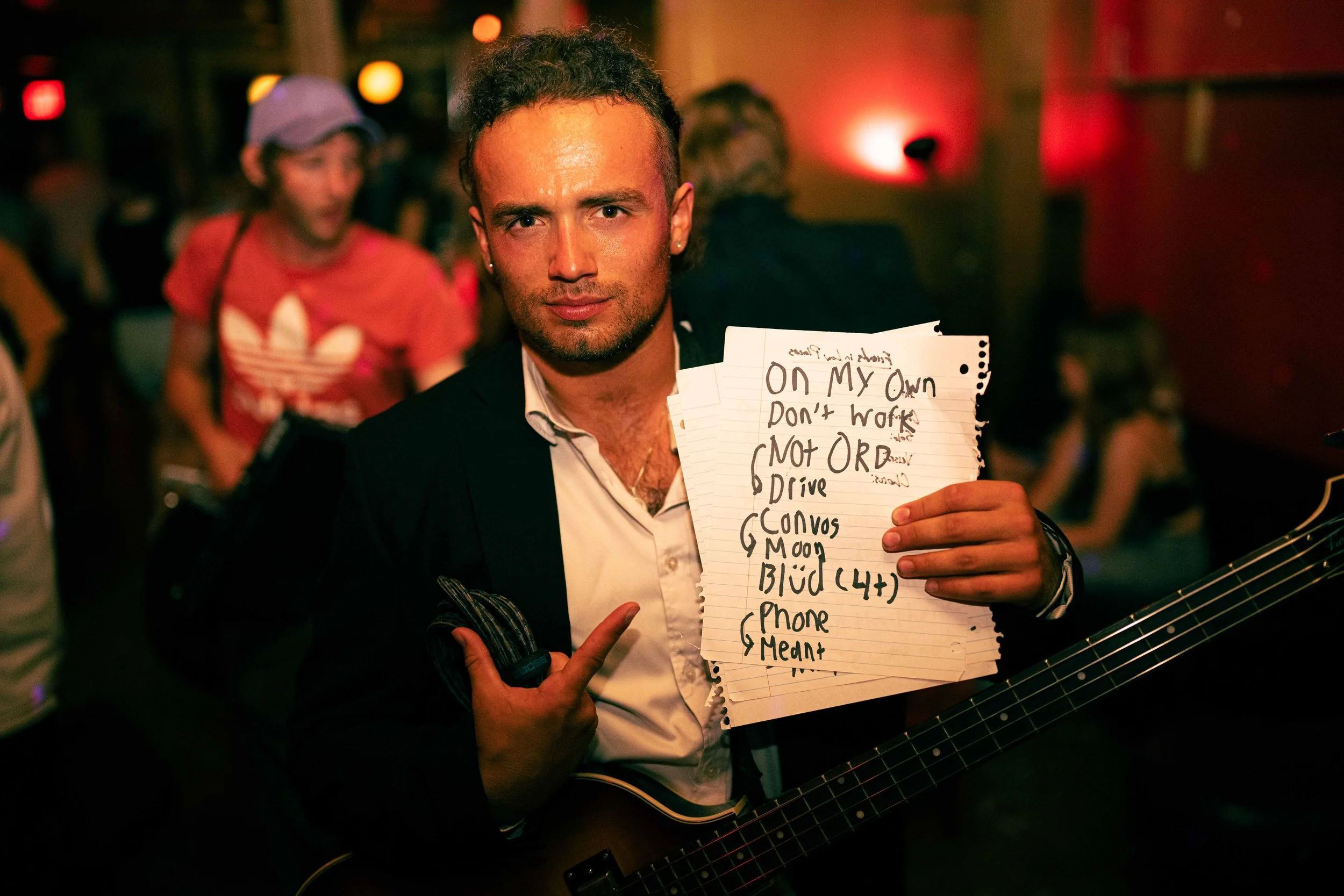 Man in suit holding a handwritten note and guitar in a dimly lit social setting.