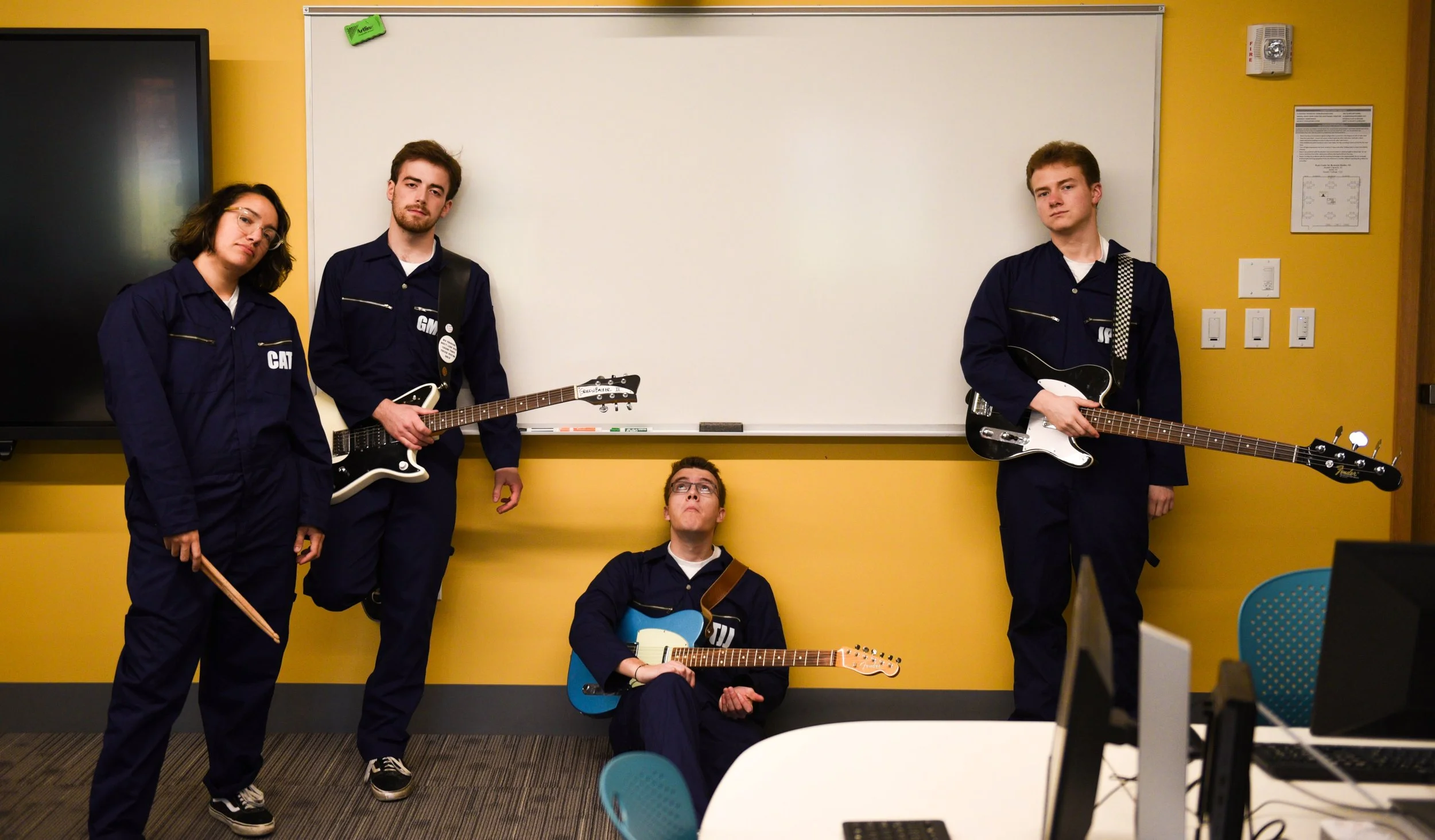 Four young individuals in navy blue jumpsuits, some holding guitars and drumsticks, pose in a room with yellow walls and a whiteboard. One person is sitting on the floor with a blue guitar, while the others stand, with one holding a different guitar 
