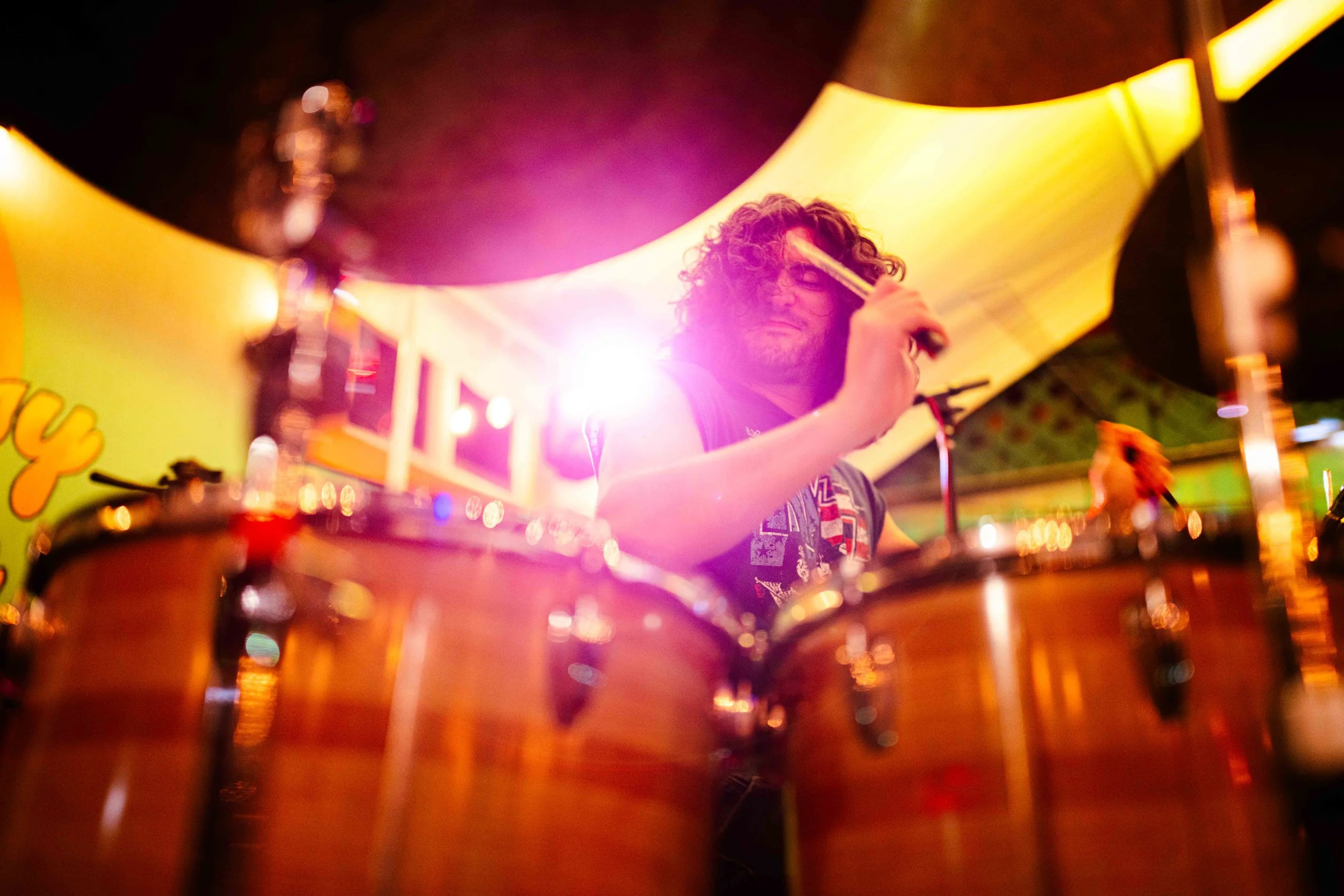A drummer with curly hair and glasses playing drums under colorful stage lighting.