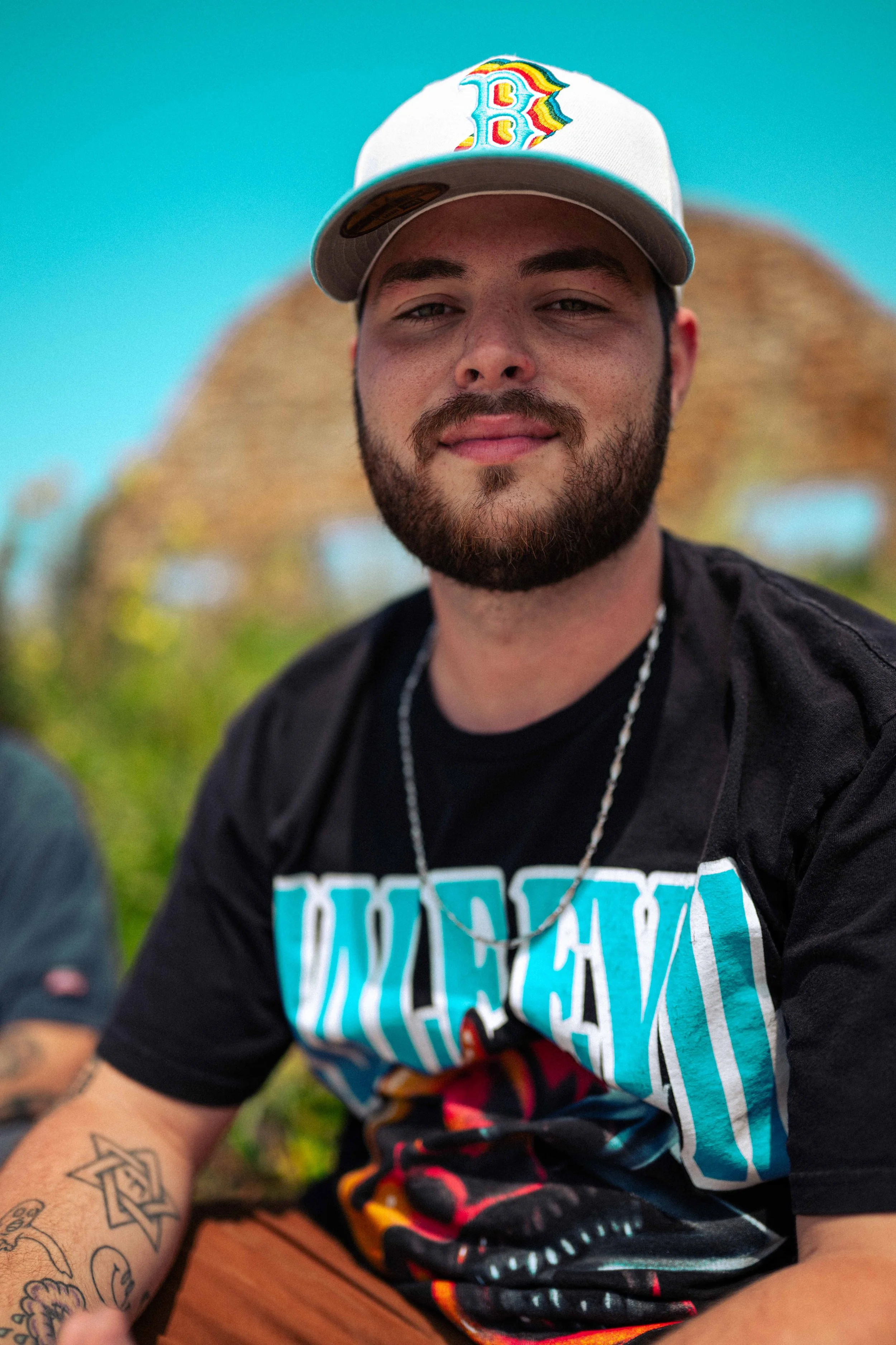 A young man with a beard wearing a white baseball cap with a colorful 'B' logo, a black T-shirt with a graphic design, and a silver chain necklace. He is outdoors with a blurred background of a blue sky and palm trees.