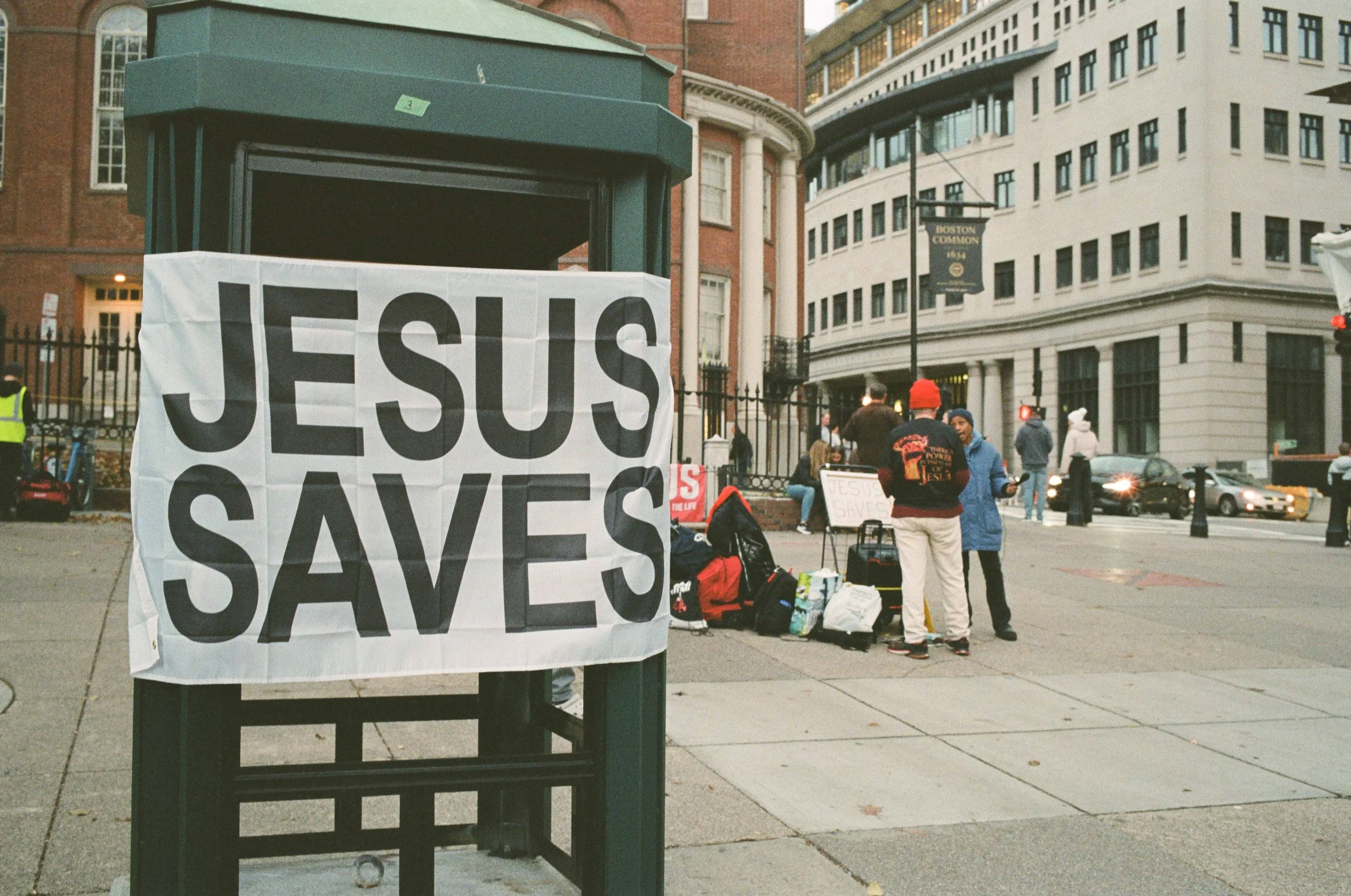 A large sign on a city street corner reads 'Jesus Saves'. In the background, people gather near some luggage and signs, with buildings and traffic in the city scene.
