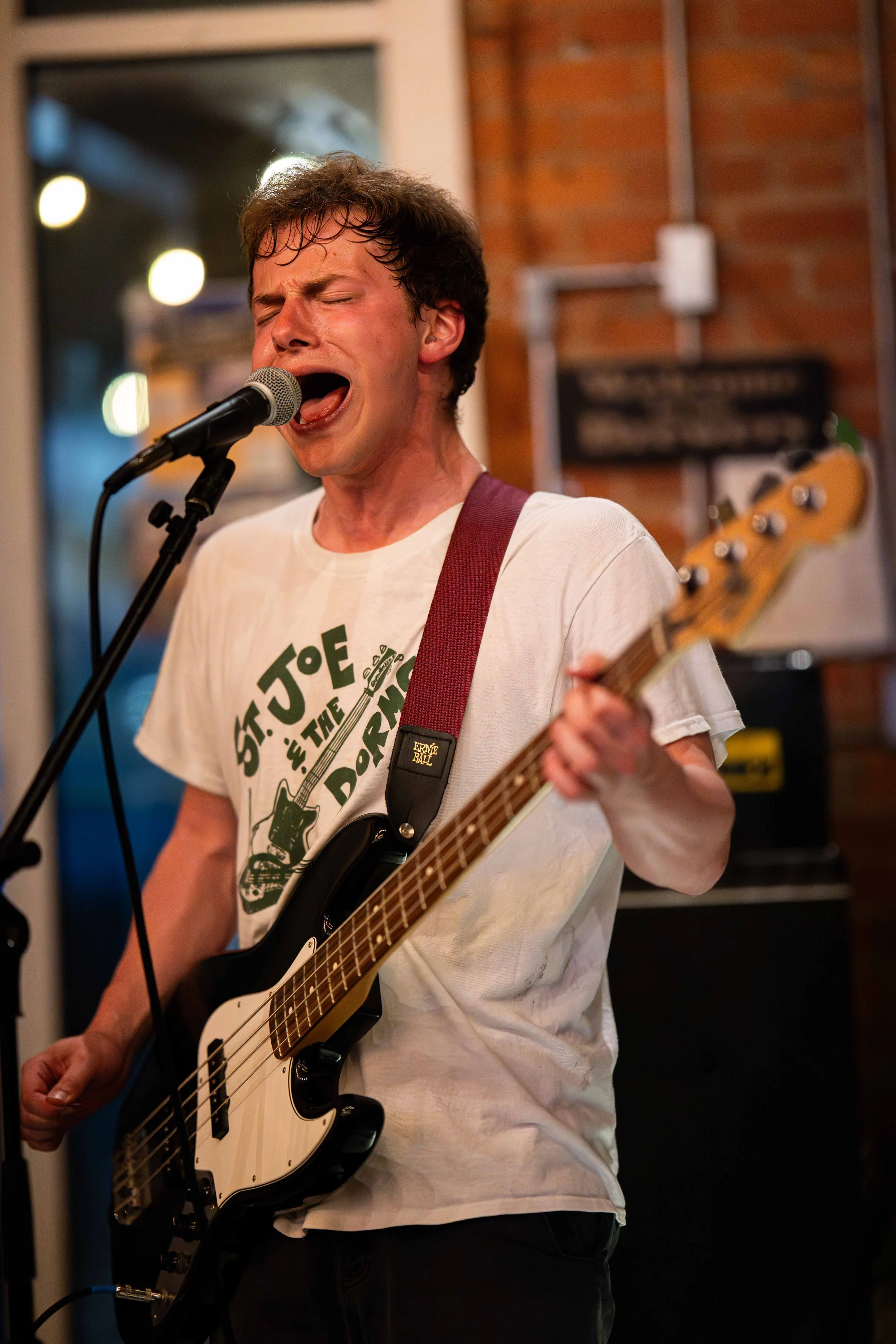 Man passionately singing into a microphone while playing a bass guitar in a cozy indoor setting with a brick wall background.