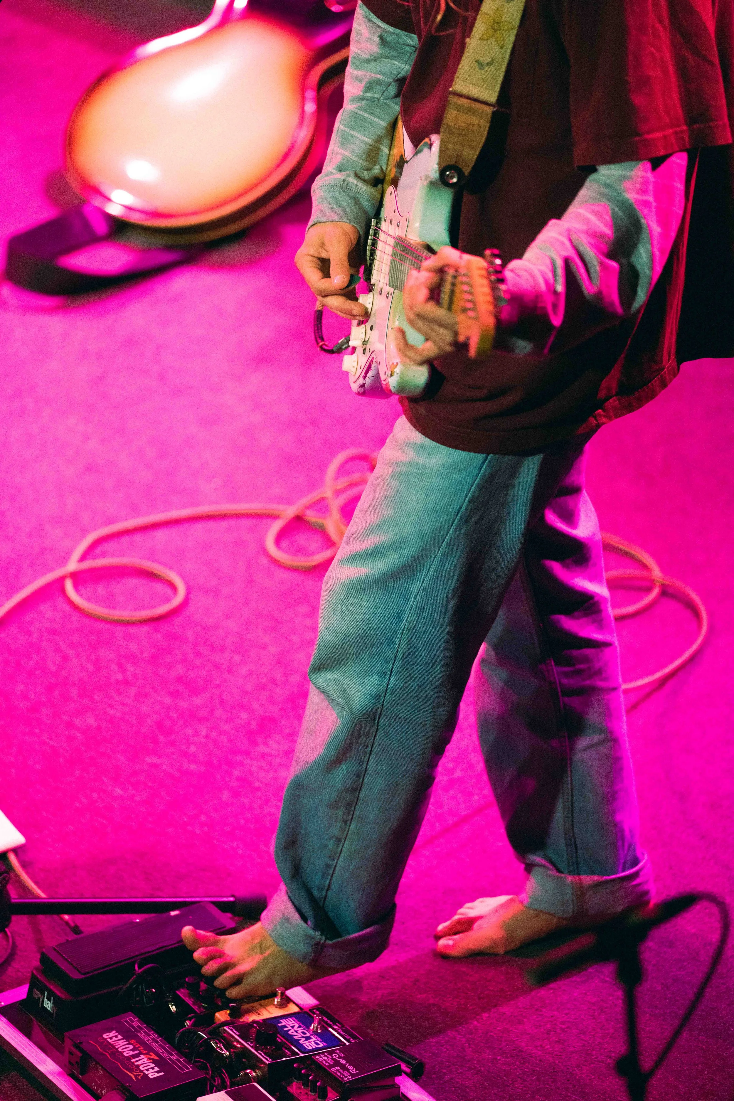 A person playing an electric guitar on stage with pink lighting, barefoot, with two guitar cases on the floor and various guitar pedals at their feet.