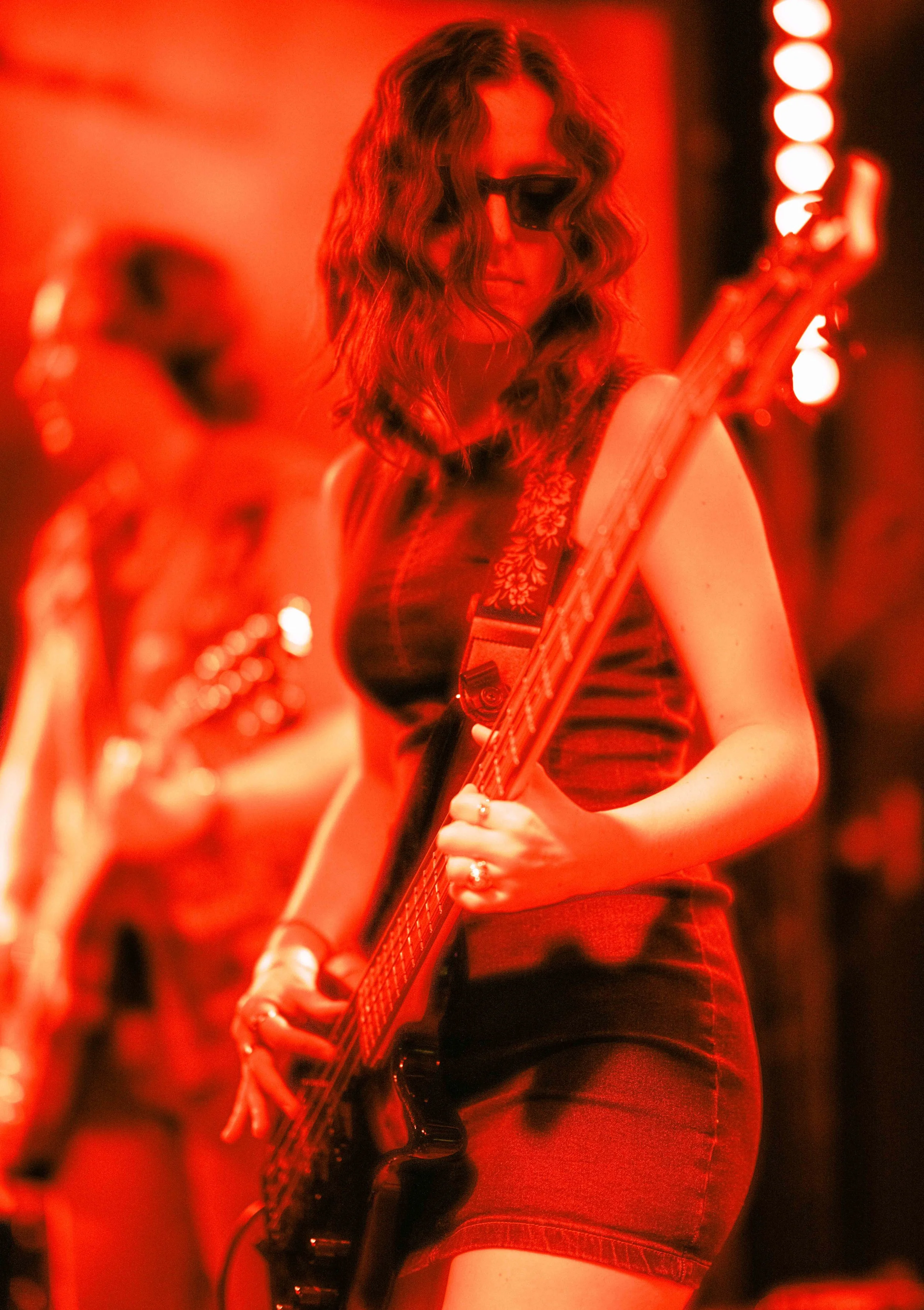A woman with curly hair wearing sunglasses and a sleeveless top playing an electric guitar on stage, with a red lighting ambiance.