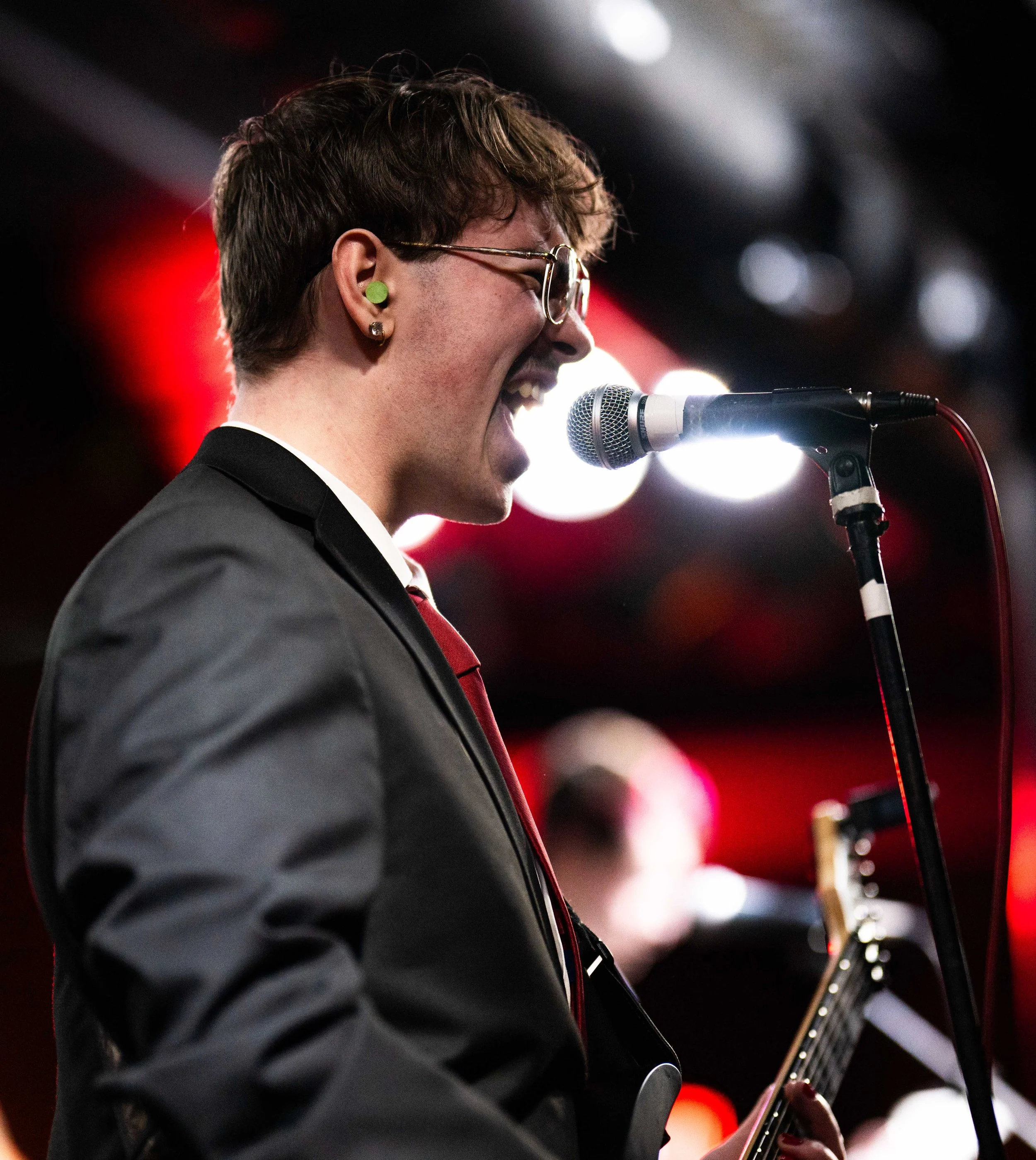 A woman singing into a microphone while playing an electric guitar on stage, wearing glasses, earrings, a suit jacket, and a tie, with colorful stage lights in the background.