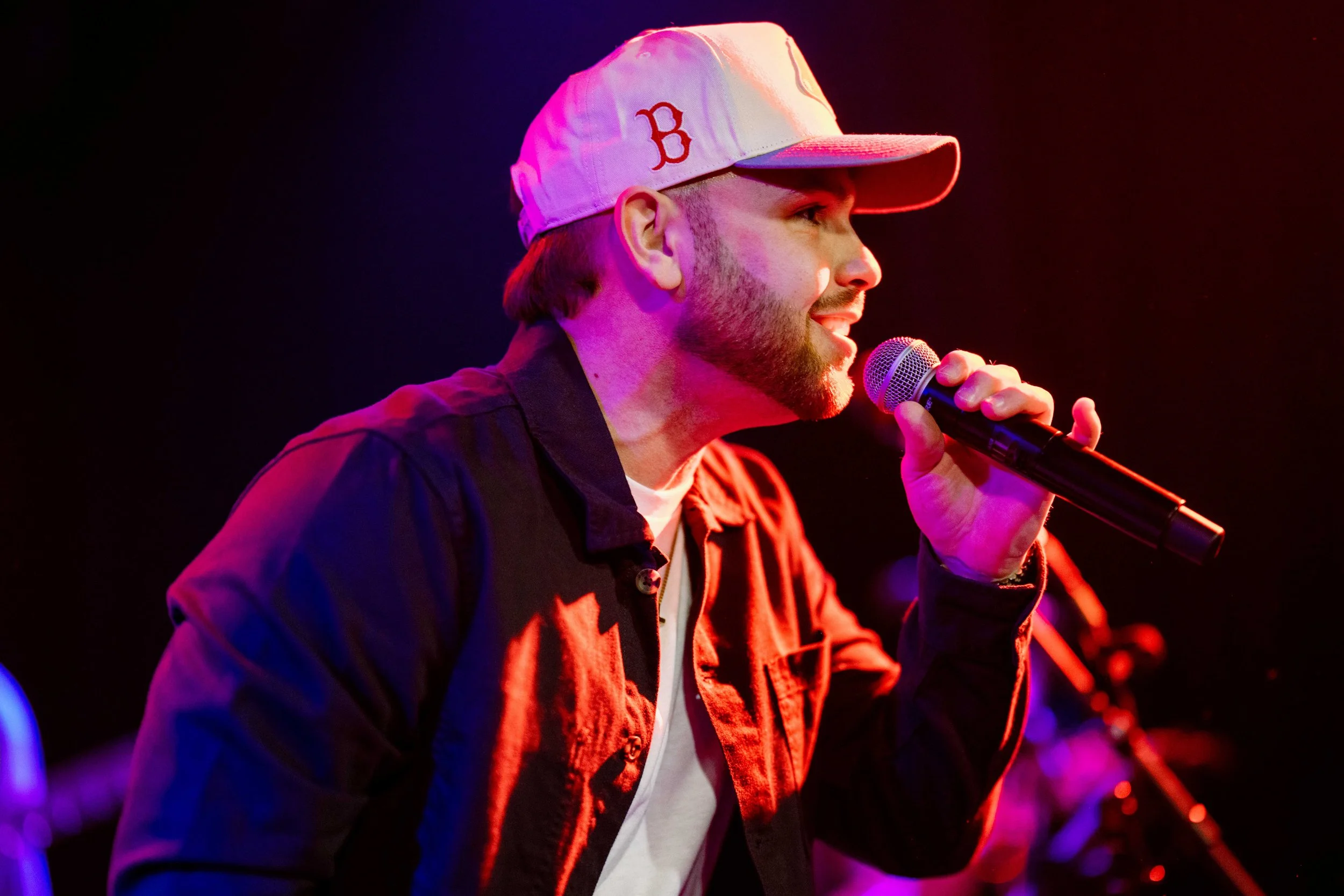 A man singing into a handheld microphone on stage, wearing a baseball cap, a dark jacket, and a white shirt, illuminated by pink and purple stage lights.