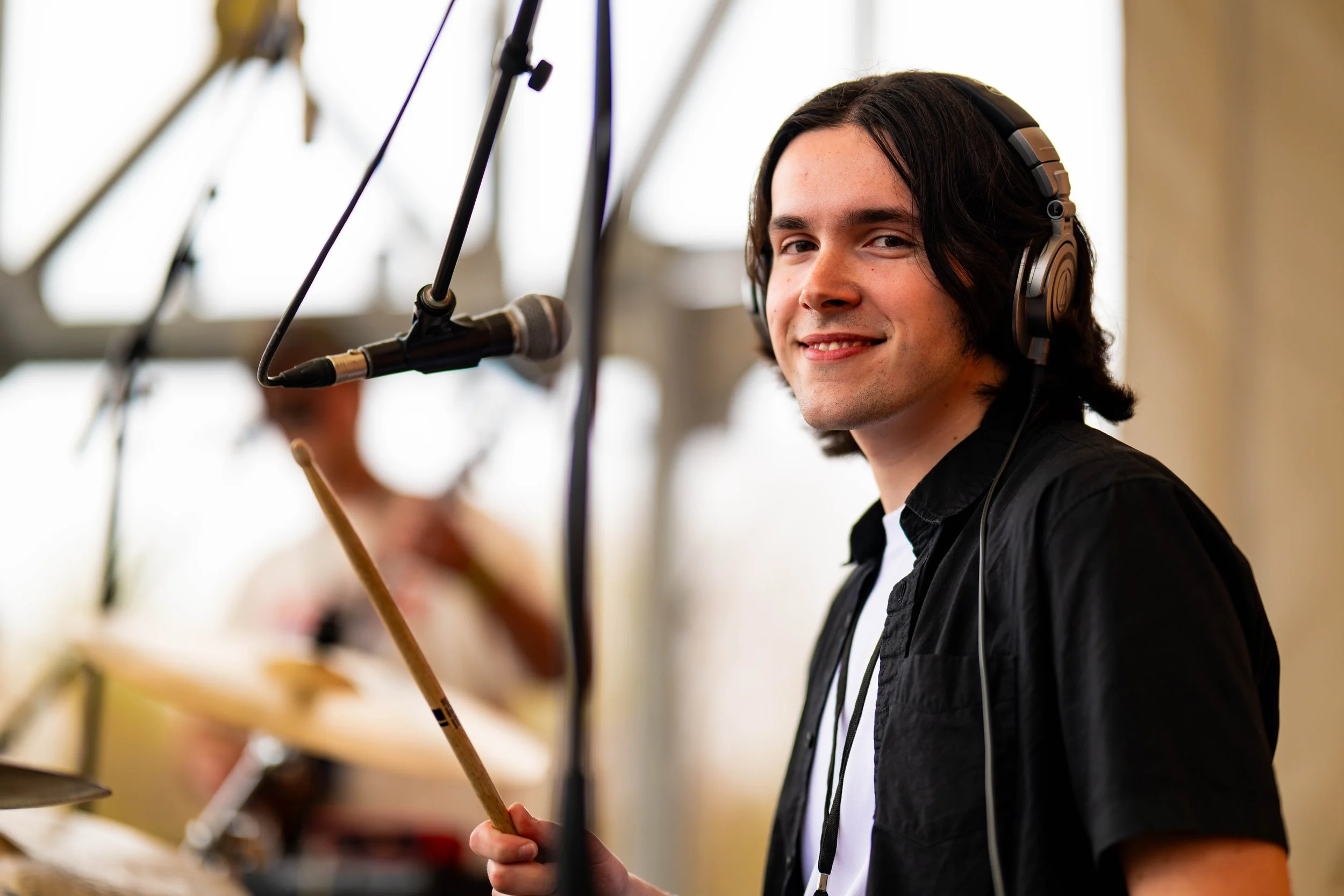 A young man with black hair wearing headphones, holding a drumstick, smiling, in a music recording studio with a microphone and a blurred background of another musician.