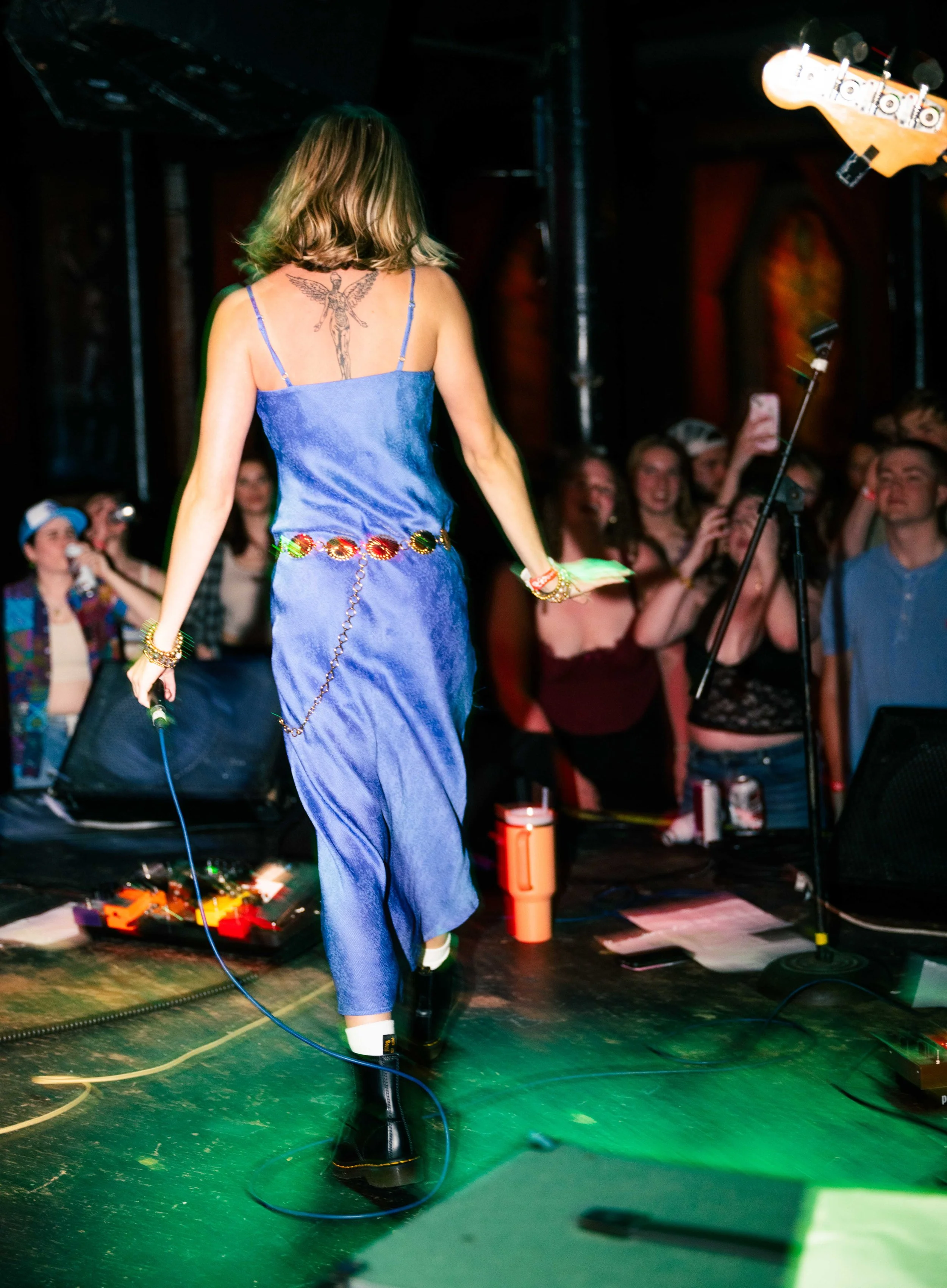 A woman on stage in a blue dress with a tattoo on her upper back, holding a microphone and facing an audience.