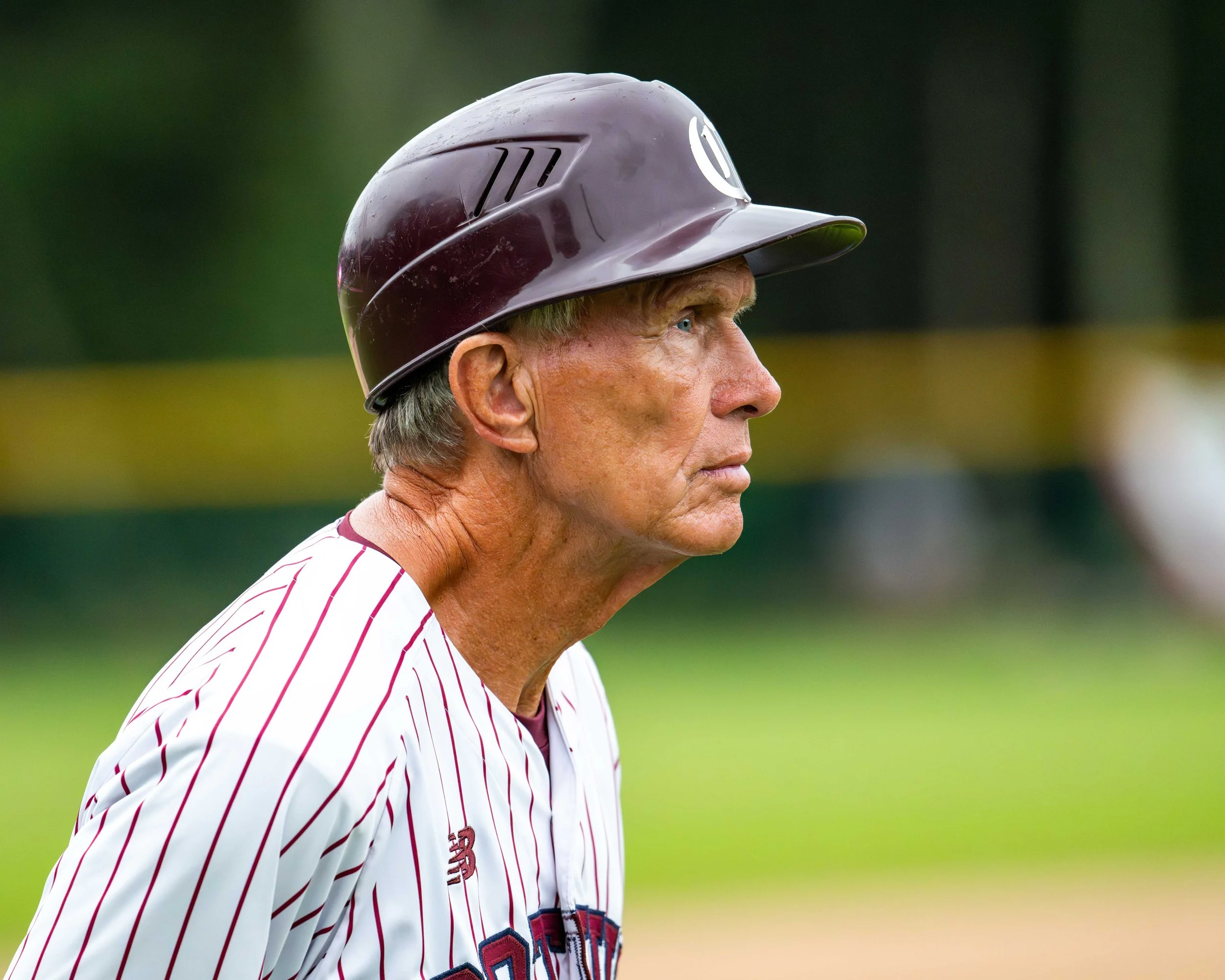 Profile of an elderly male baseball player wearing a maroon helmet and a white pinstripe uniform with red and blue accents, standing outdoors.