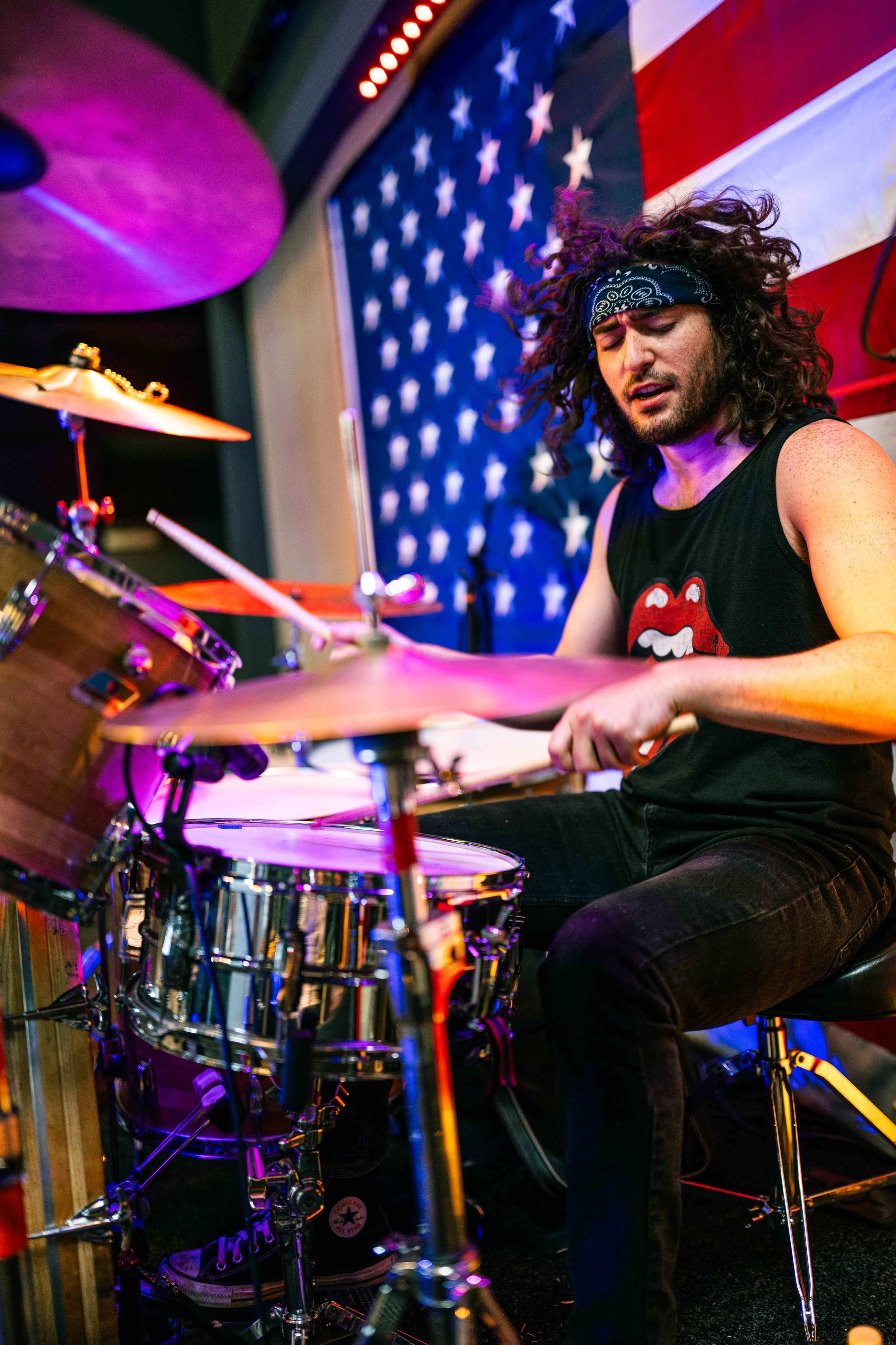 A drummer with curly hair, wearing a bandana and a black sleeveless shirt with a Rolling Stones logo, playing drums on stage with an American flag in the background.