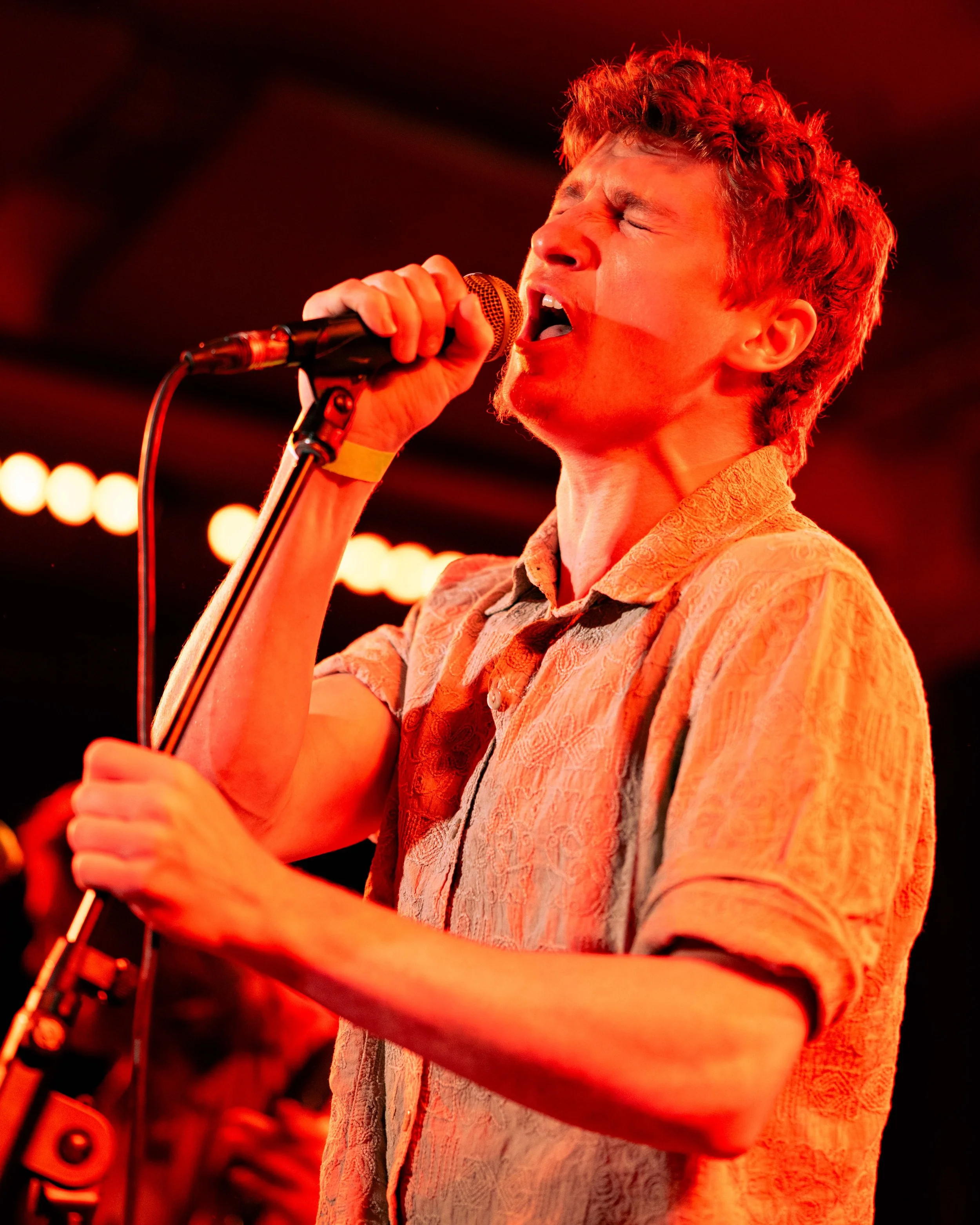 A young man singing passionately into a microphone on stage with red lighting.