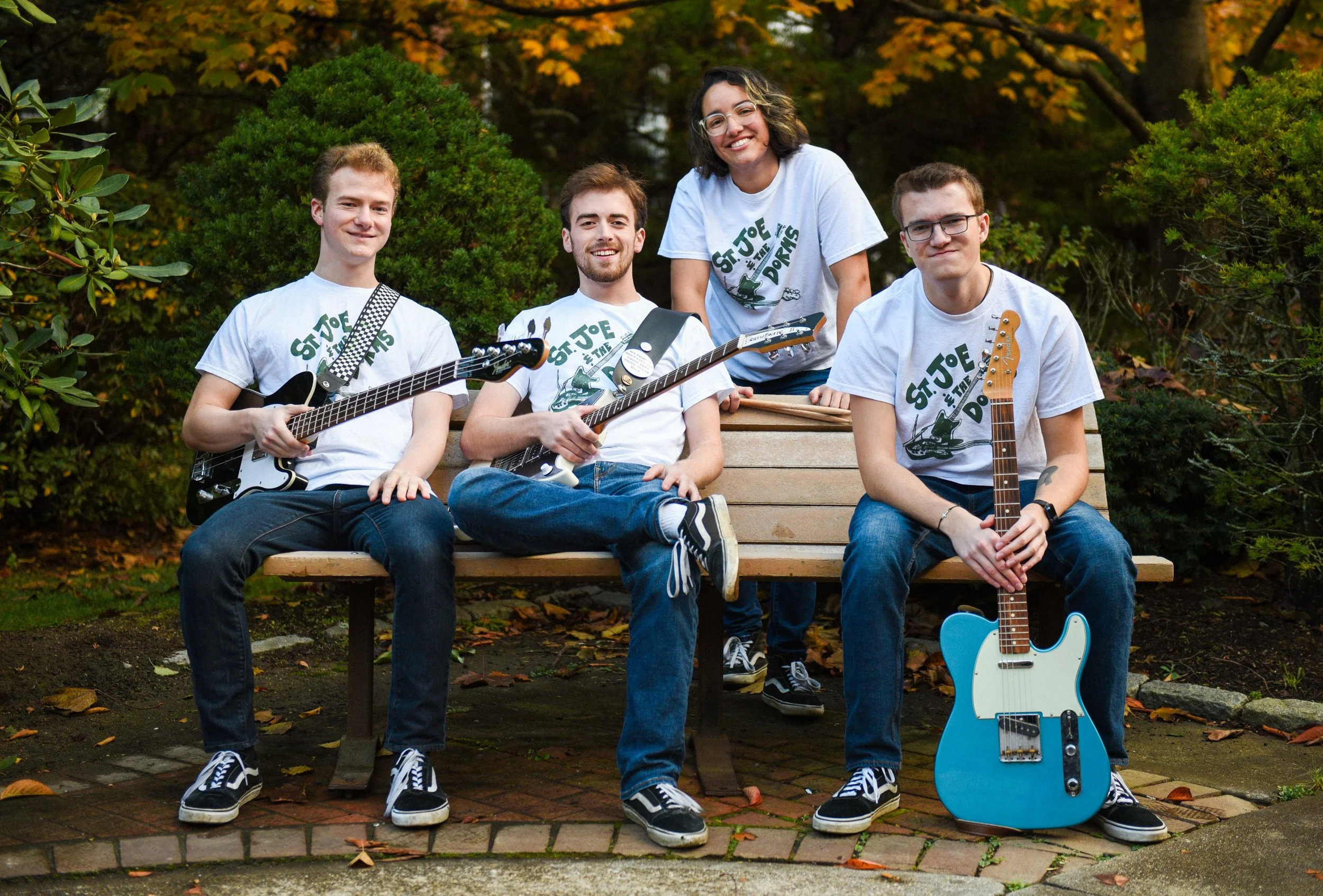 A group of four young adults, three men and one woman, sitting on a park bench outdoors, holding guitars and smiling. They are wearing white T-shirts with a guitar graphic and the text 'St. Joe in the D.' The background includes trees with autumn fol