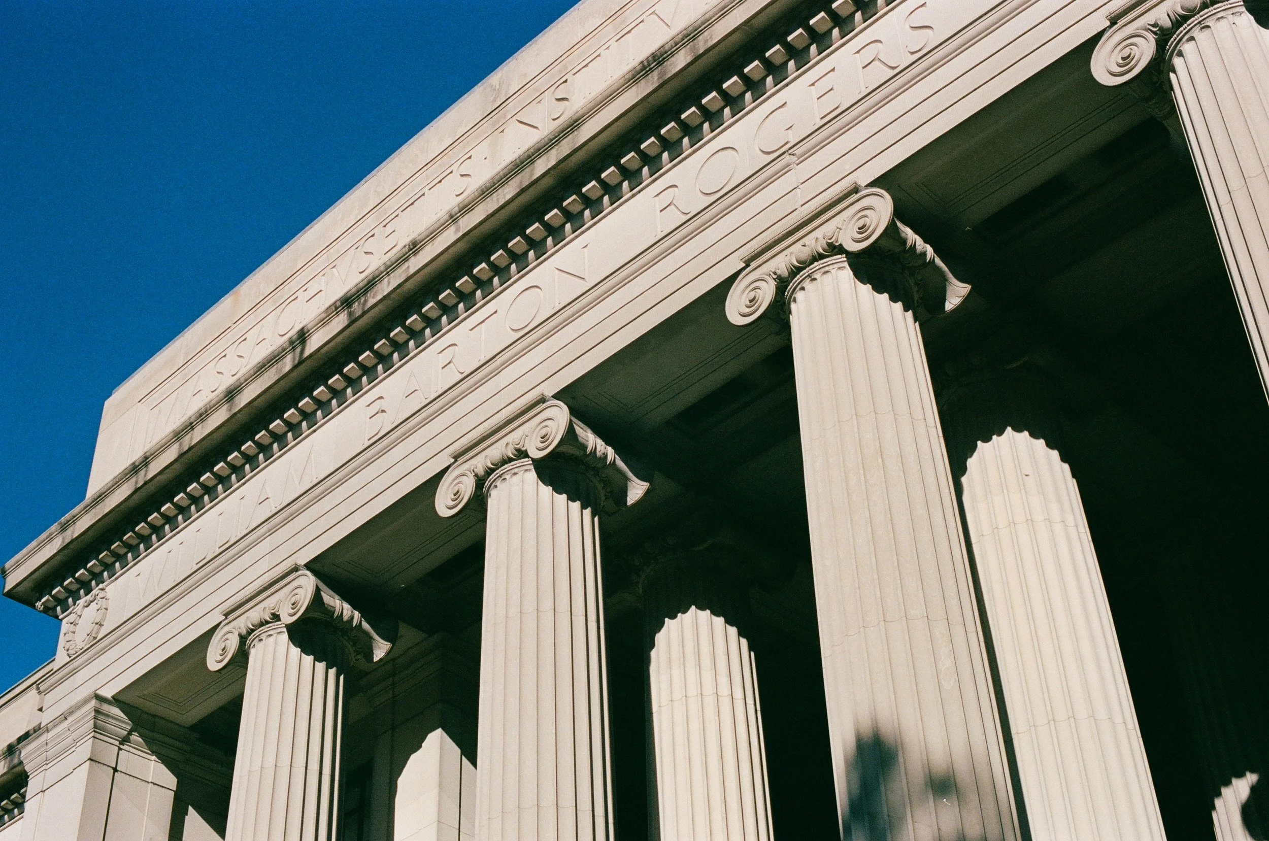 Close-up of a classical building facade with large fluted columns, decorative capitals, and inscriptions in Latin on the entablature against a clear blue sky.