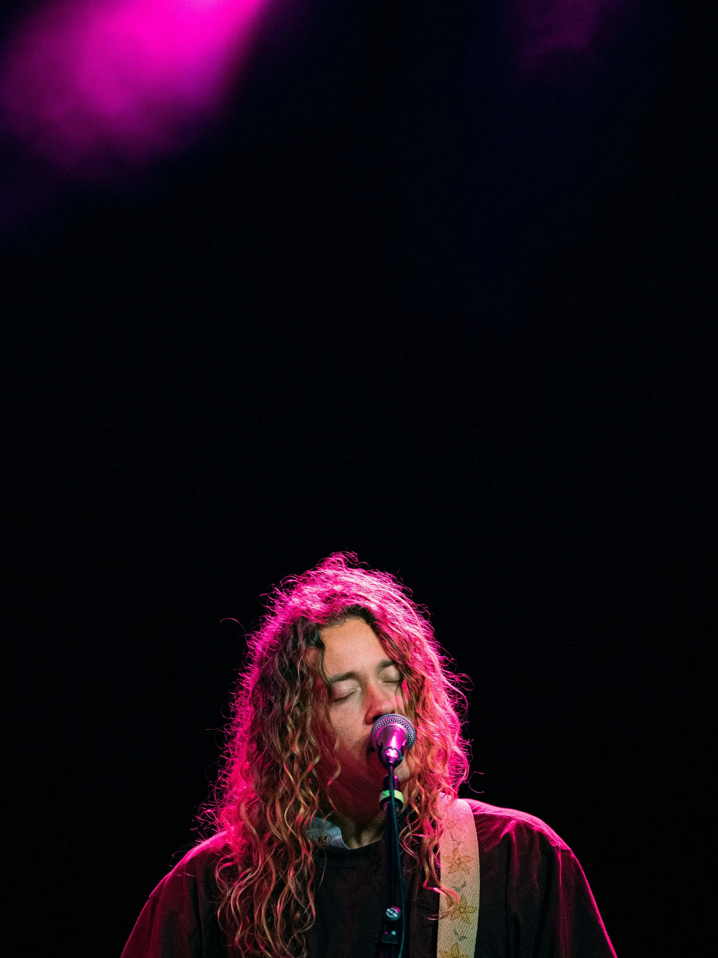 A woman with curly hair singing into a microphone on stage with pink and purple lighting.