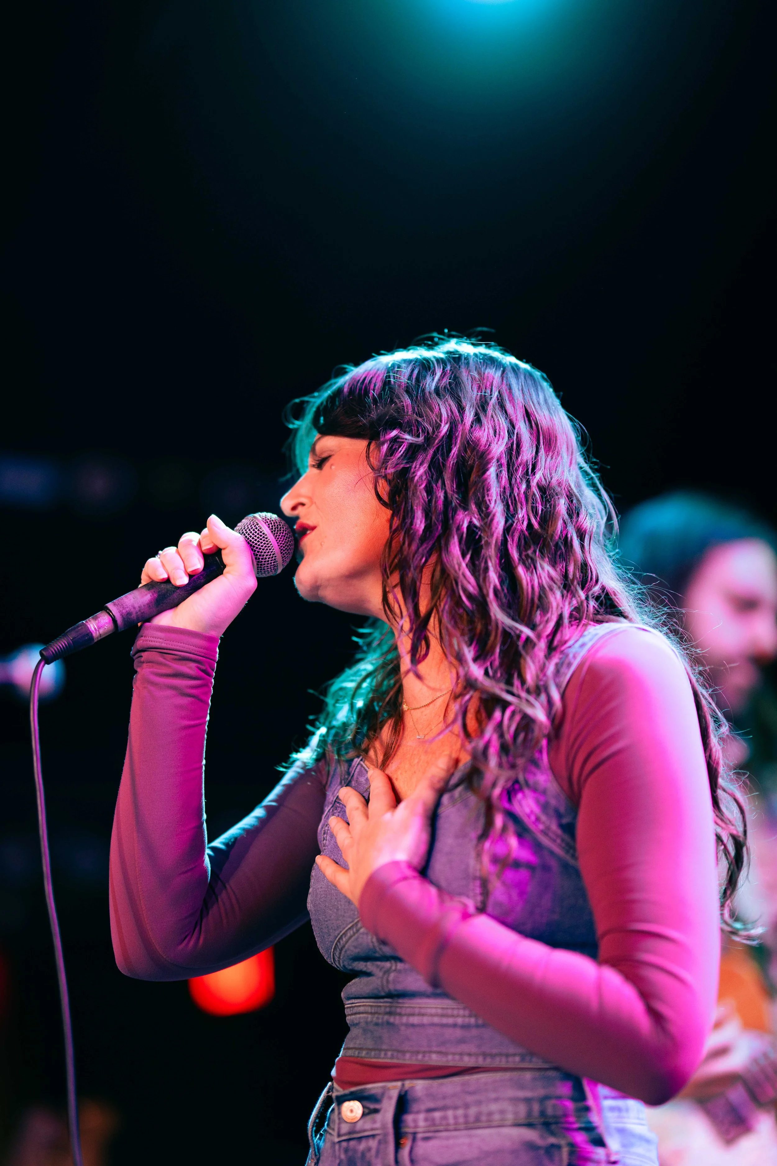 Woman singing on stage with colorful lighting, holding microphone with one hand and placing her other hand on her chest.