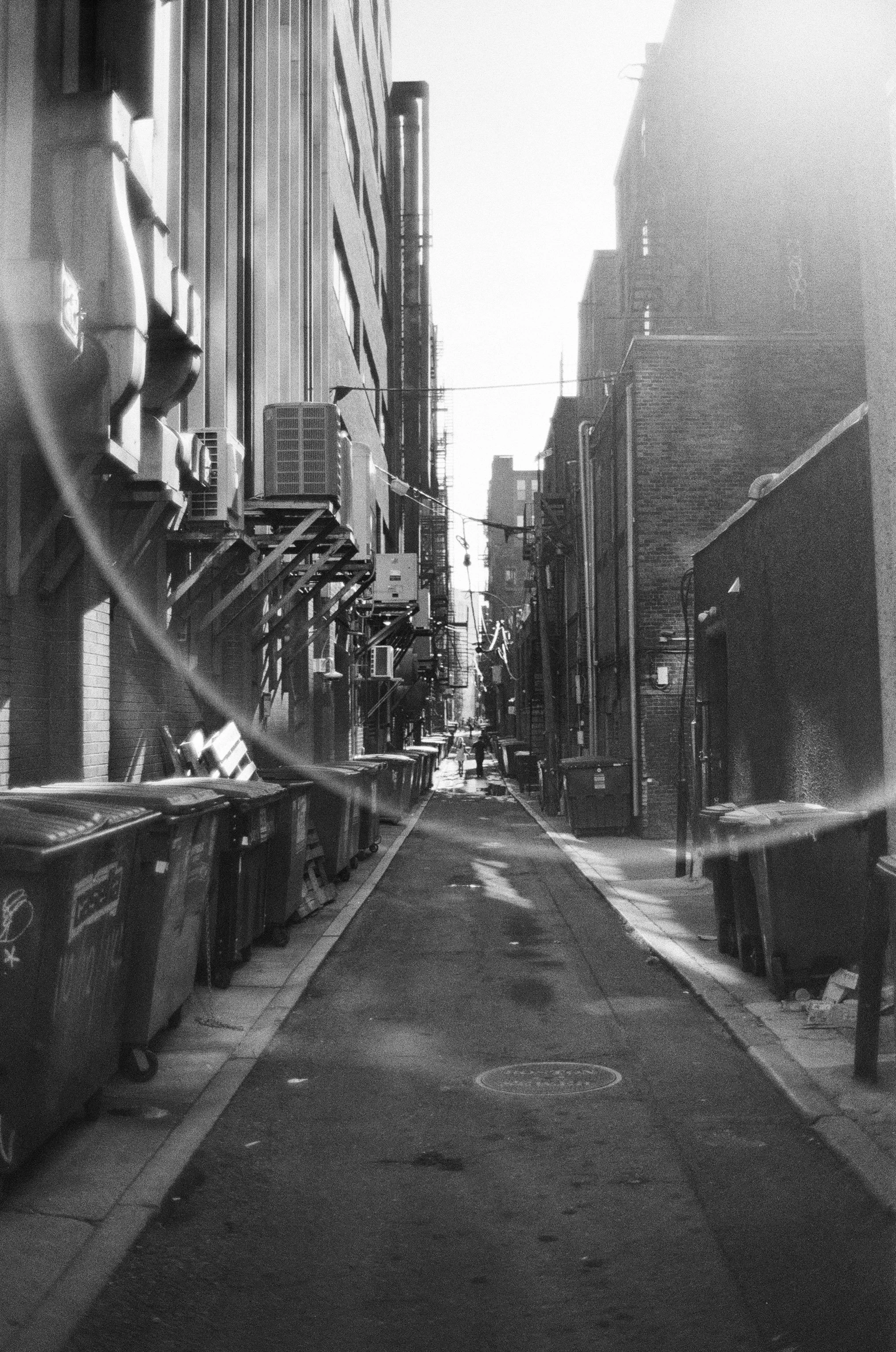 Black and white photograph of an alleyway lined with trash bins, buildings, and visible power lines overhead, with a few people walking in the distance.