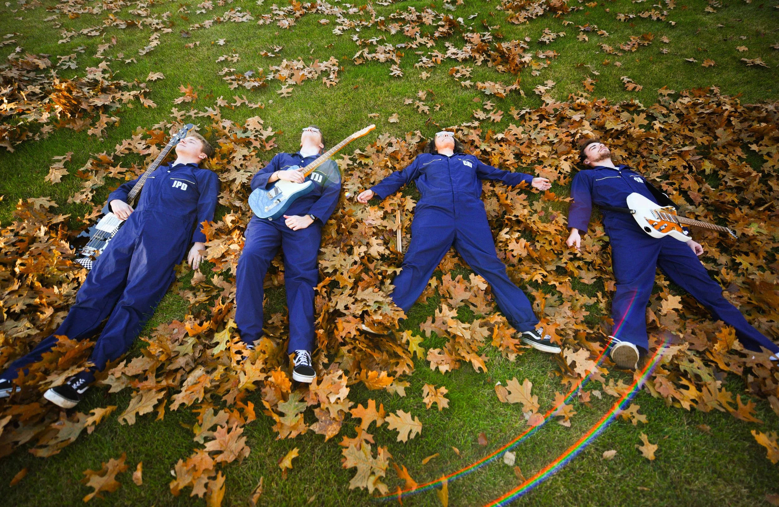 Four people lying on the grass surrounded by autumn leaves, holding guitars, and relaxing with their eyes closed.