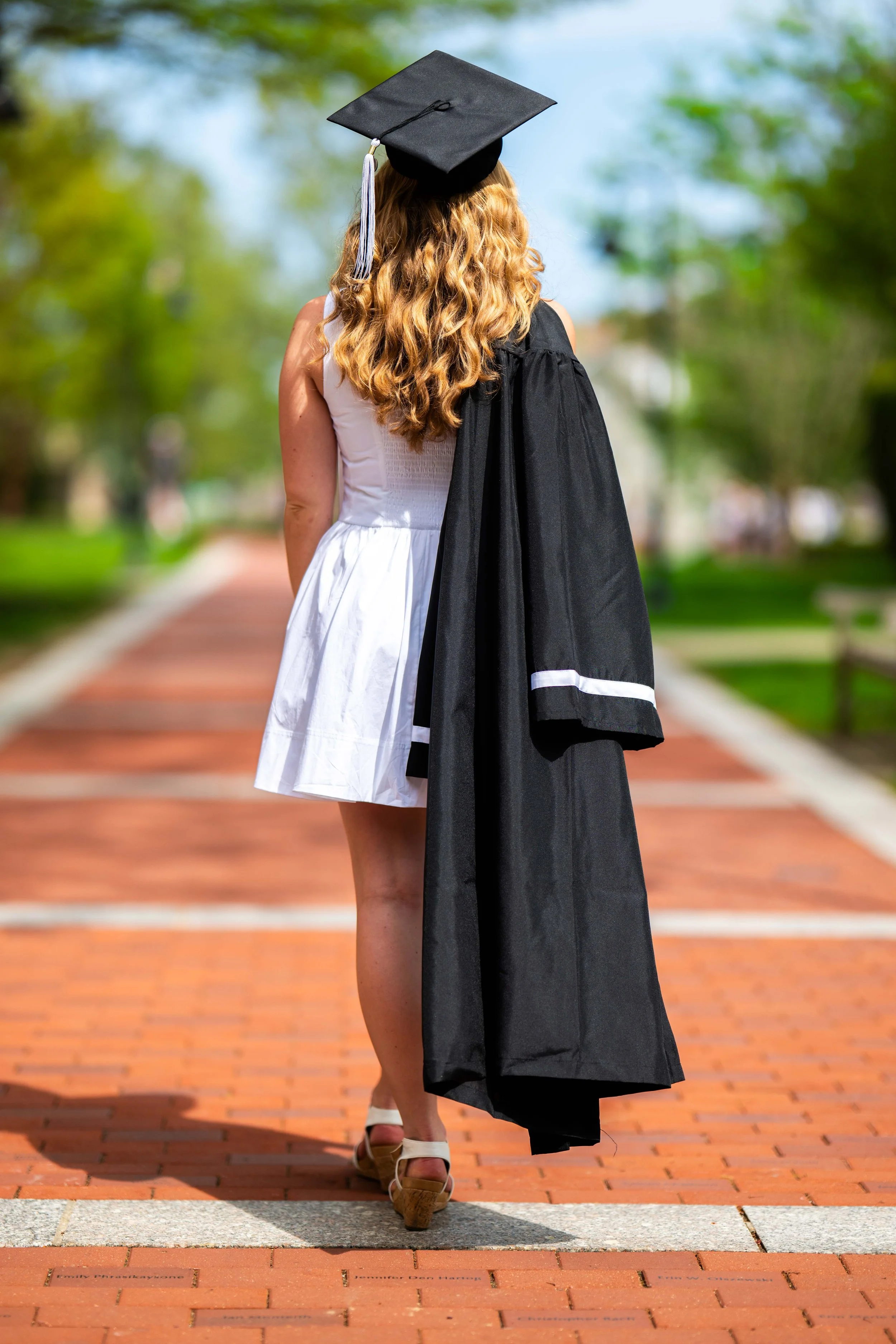 A woman in a white dress is walking on a brick pathway outdoors, wearing a graduation cap and holding a graduation gown over her shoulder.