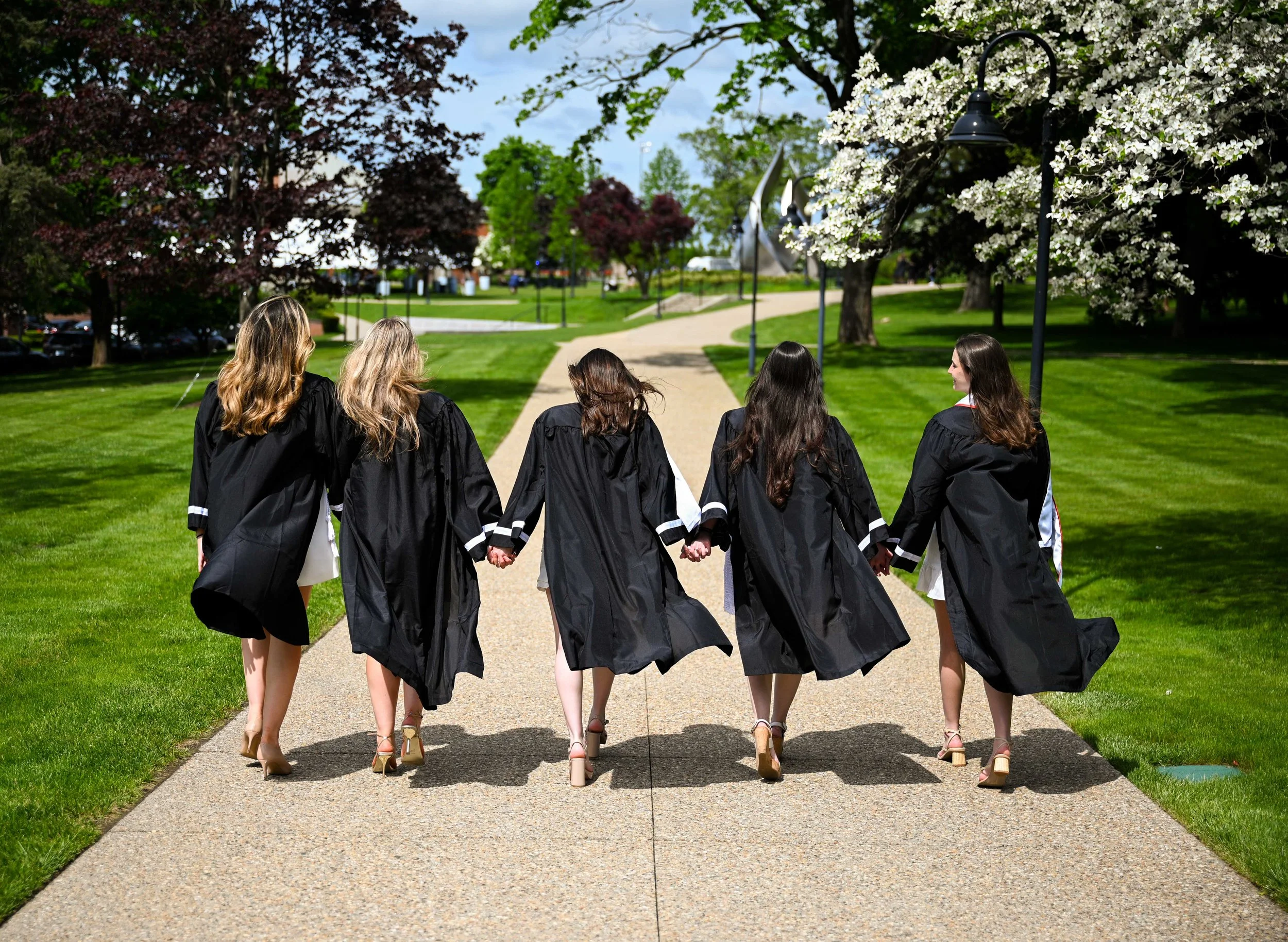 Group of five women in graduation robes walking hand-in-hand on a paved path in a park with trees and blooming flowers.