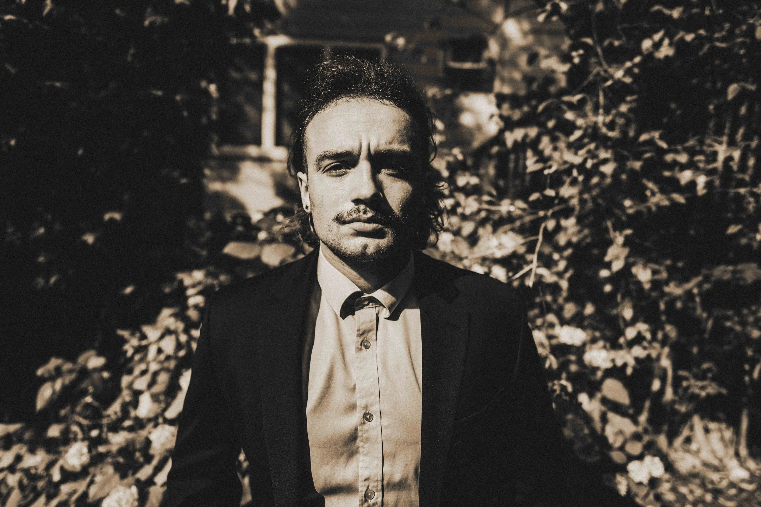 A black and white photo of a man with dark hair, a mustache, and a goatee, wearing a suit and standing outdoors with a background of trees and a house.