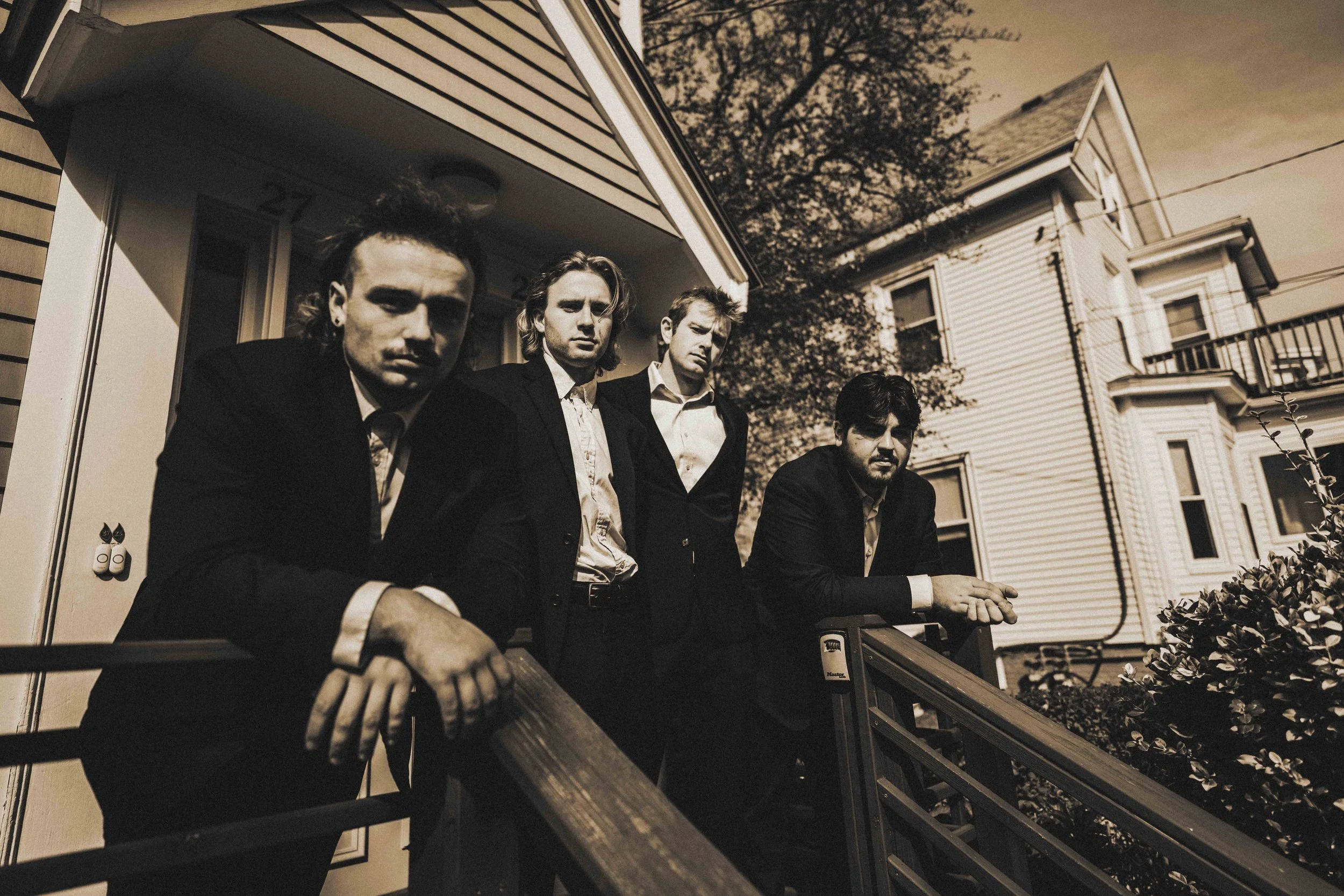 Four men in suits standing on a porch in front of a house, looking at the camera. The photo has a sepia tone.
