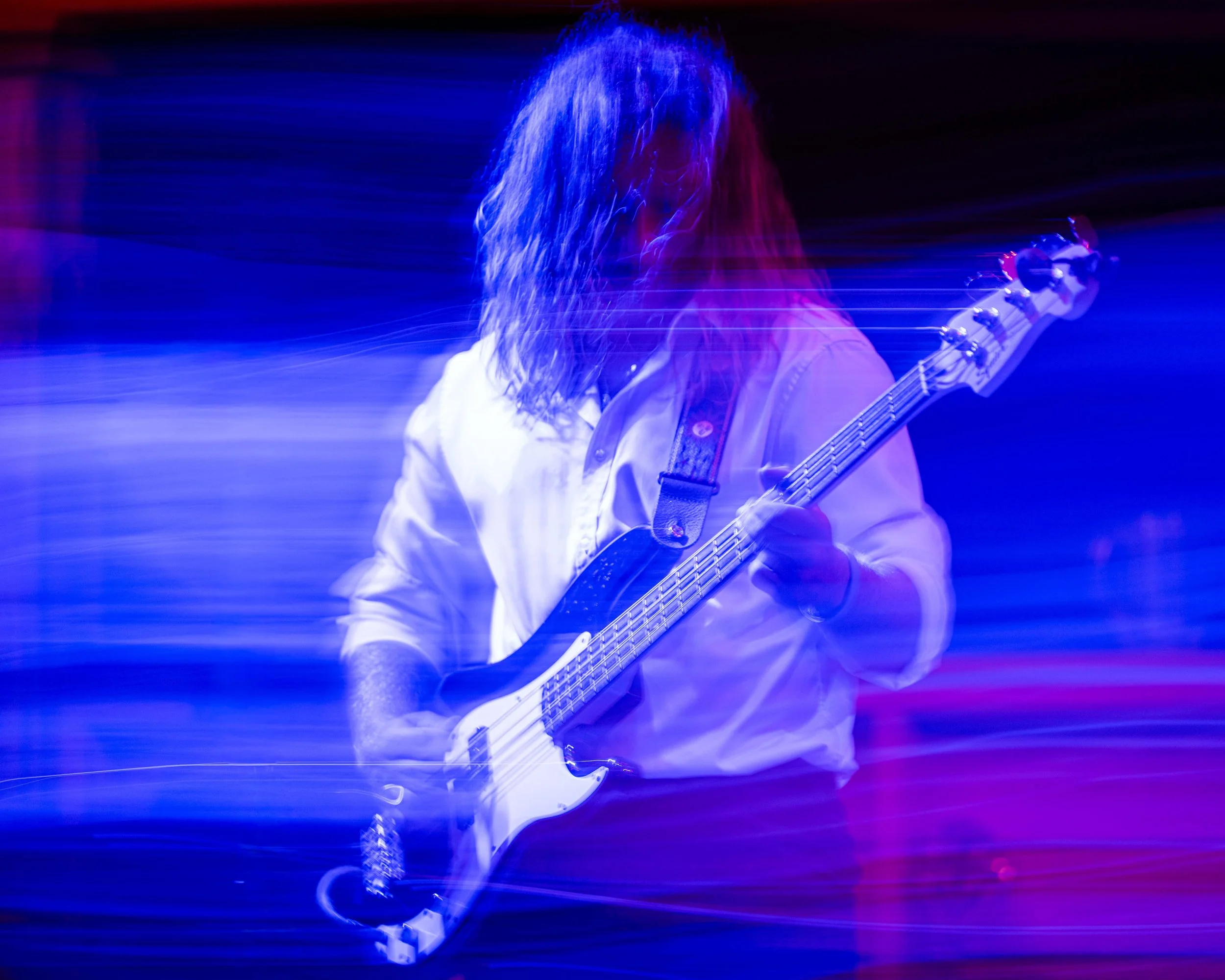 A person playing an electric bass guitar on stage under colorful, moving lights, with their hair obscuring their face.