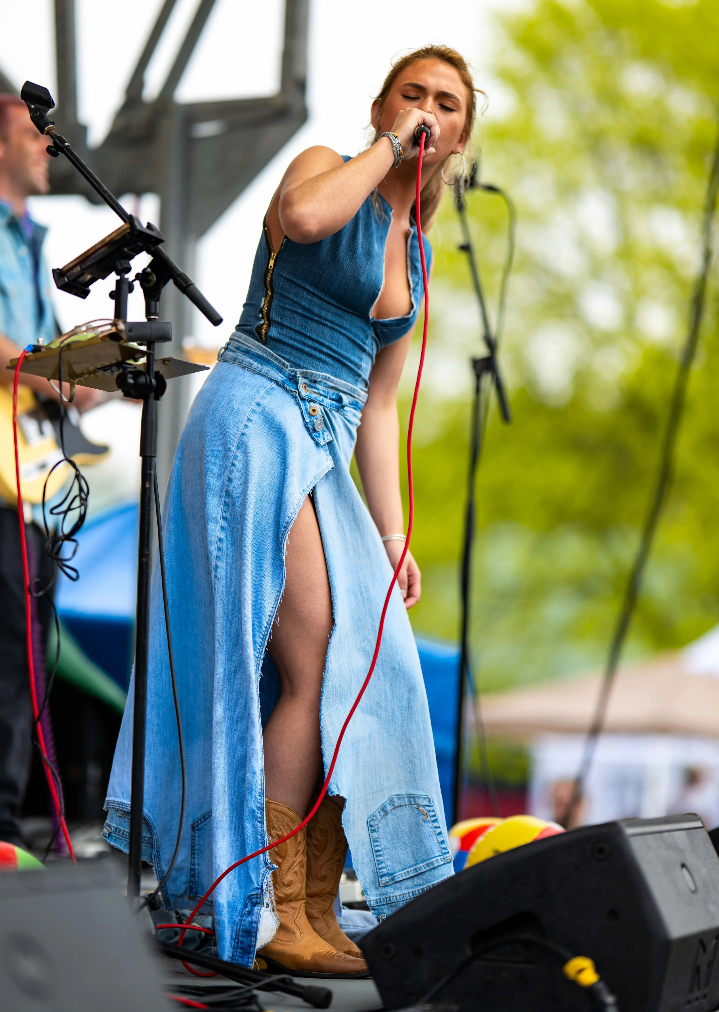 A woman singing into a microphone during a live outdoor music performance, with a musician playing guitar in the background.