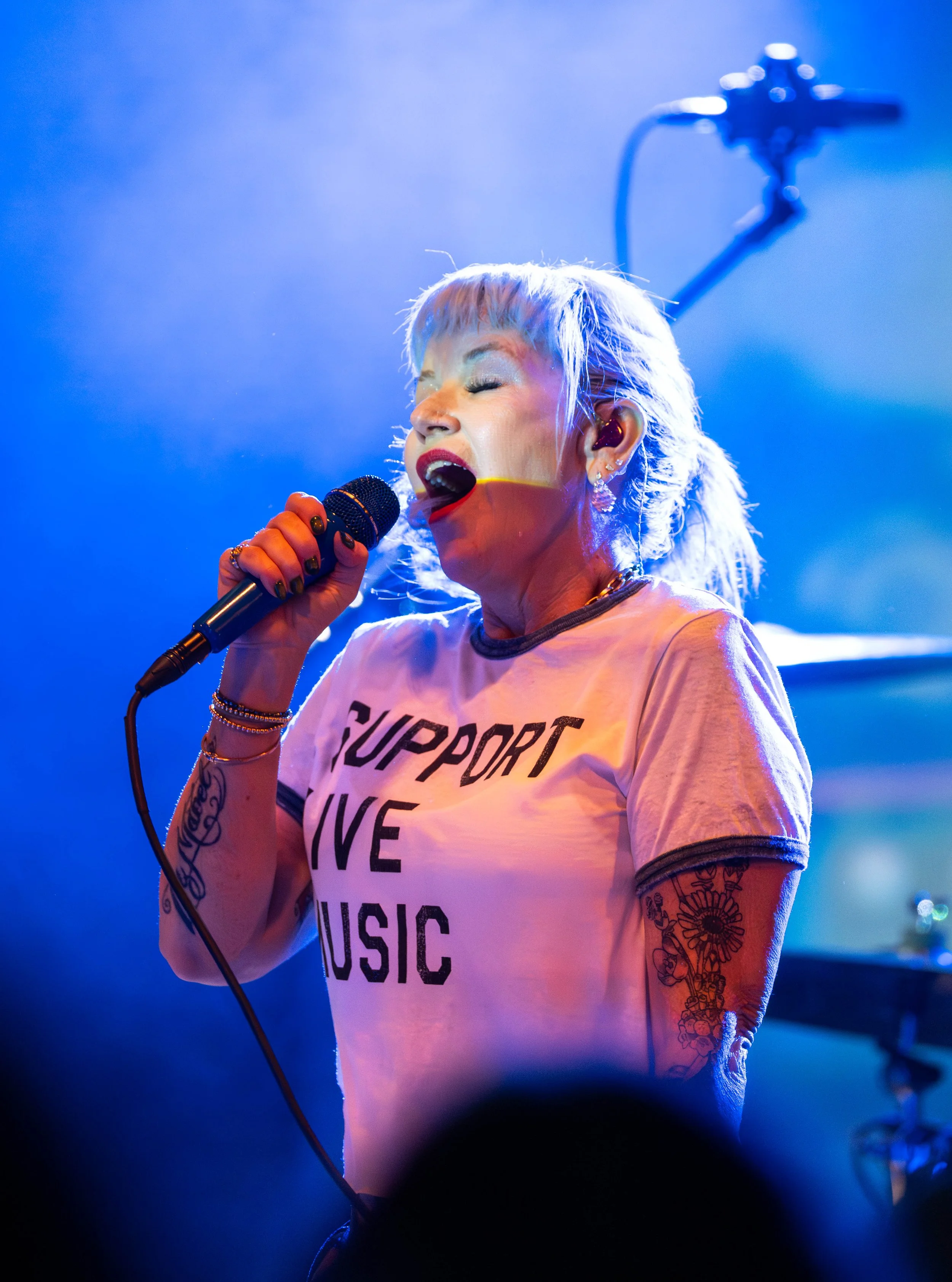 A woman singing passionately into a microphone on stage, with blue lighting and musical equipment in the background.