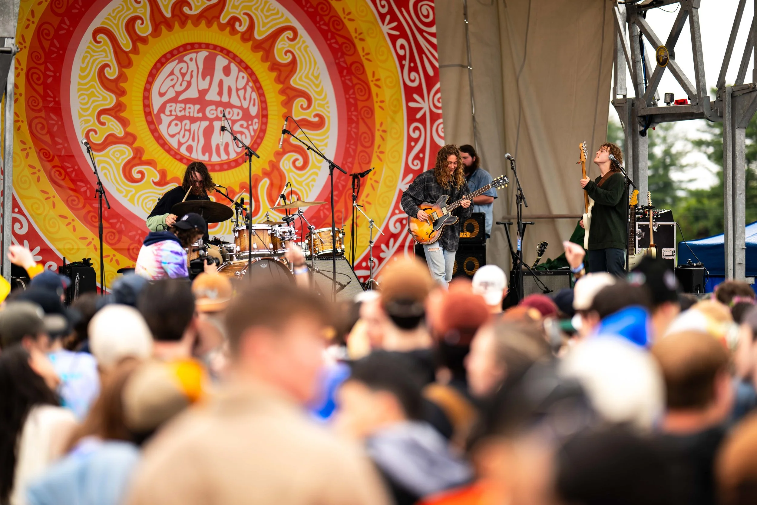 A band performing live on a stage at an outdoor music festival with a colorful circular backdrop that has a sun design and the text 'REALLY REAL GOOD OUT' and some person in the audience watching.
