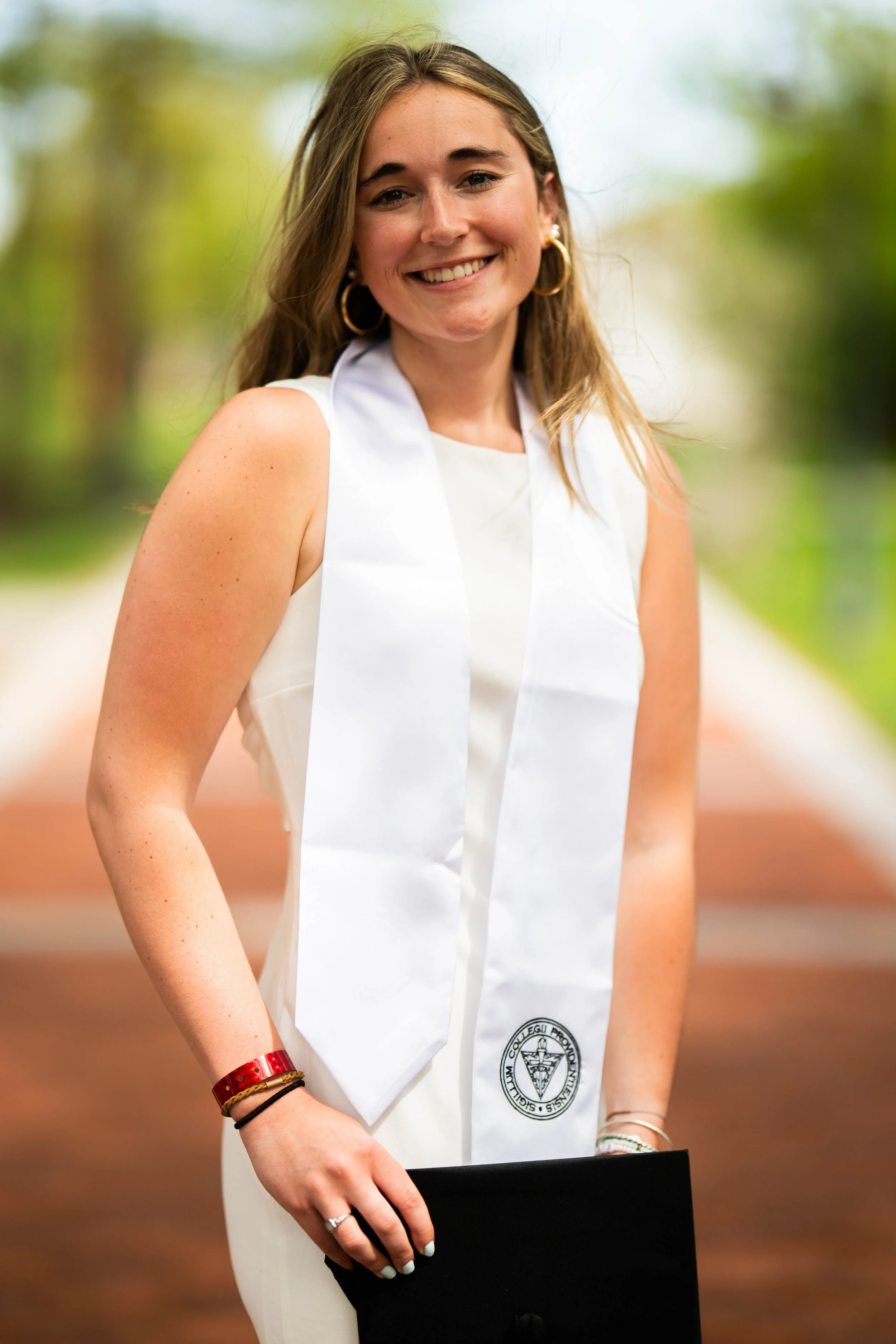 Young woman smiling outdoors in a white dress with a white graduation stole, holding a black diploma folder, wearing gold hoop earrings and a red bracelet.