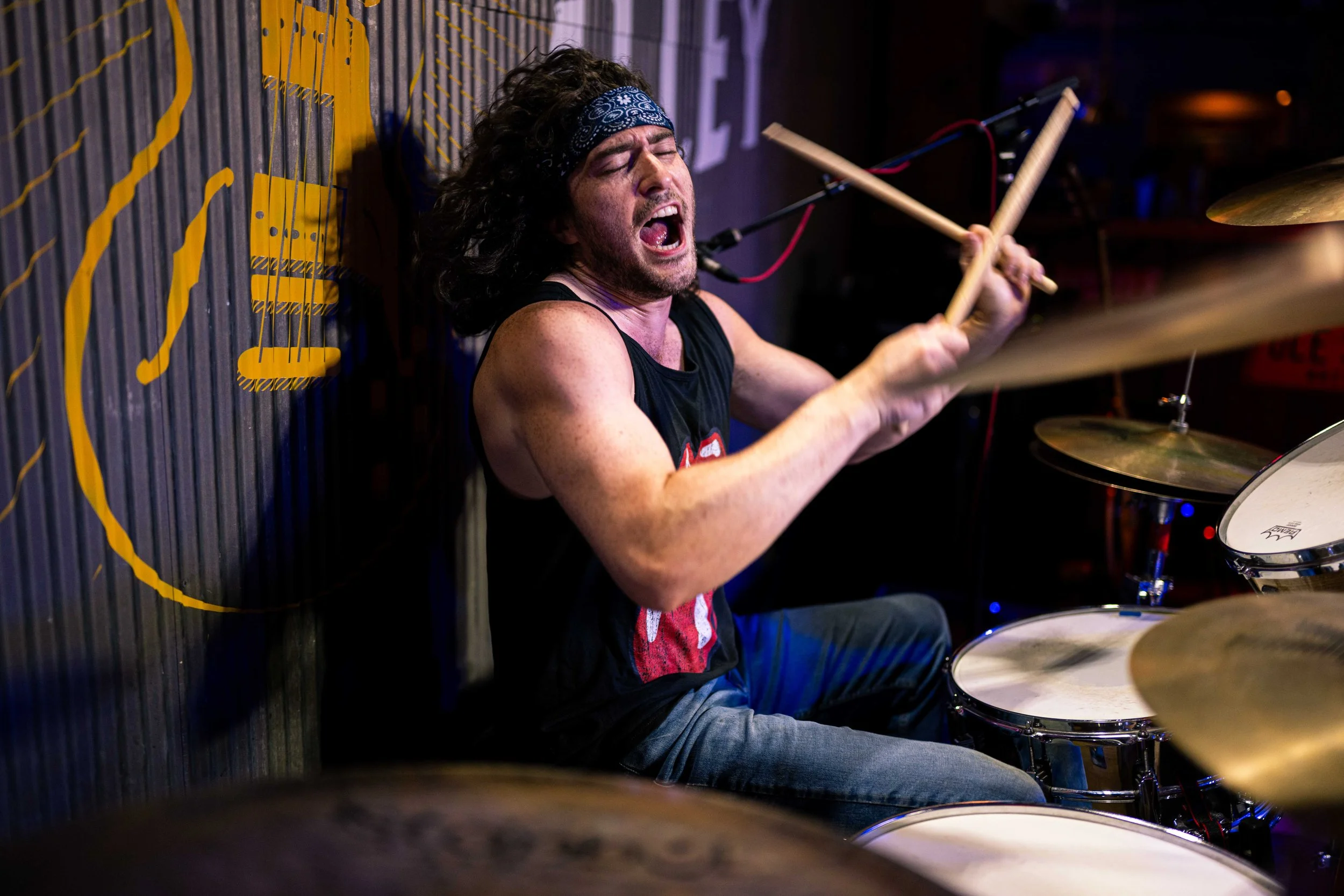 A male drummer with long curly hair, a blue bandana, and a sleeveless black shirt passionately playing drums during a live performance.