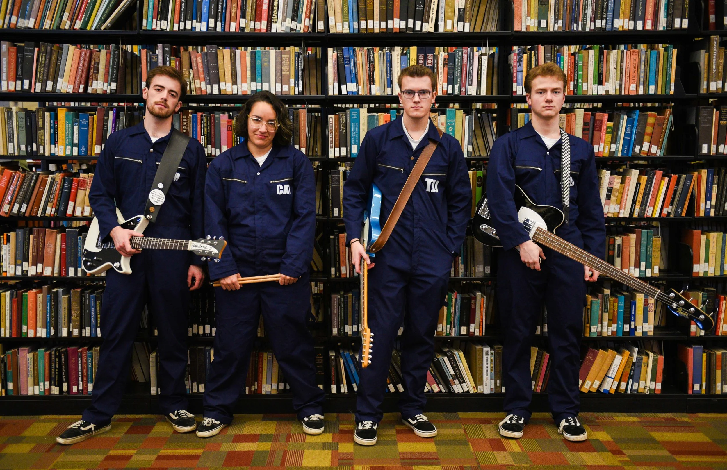 A group of four young people standing in front of a bookshelf in a library, each holding a musical instrument, all wearing blue jumpsuits.